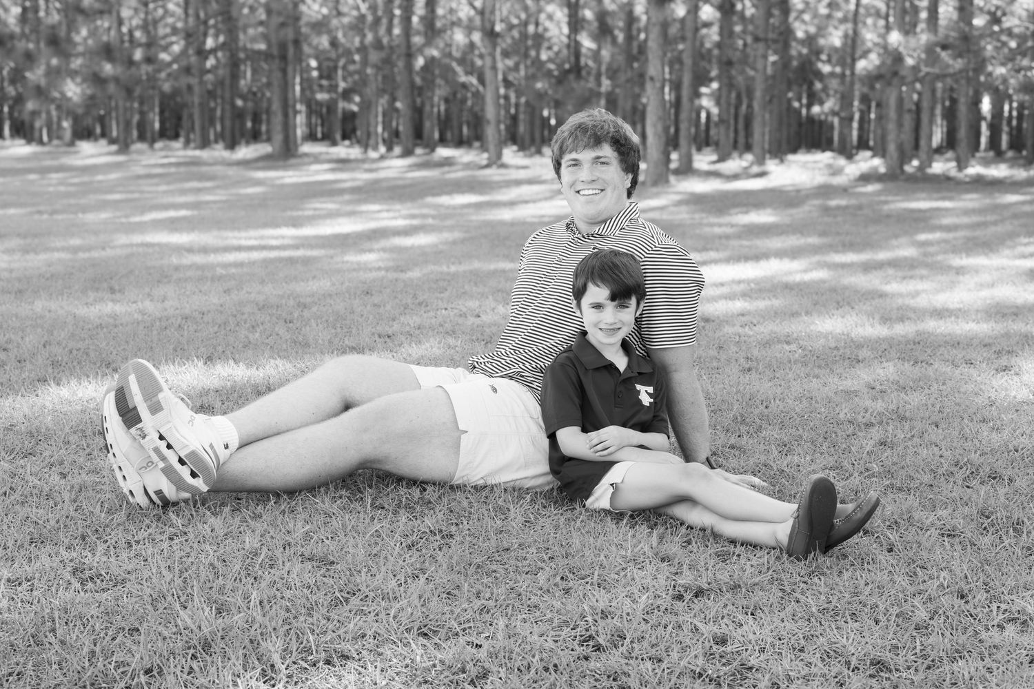 Black and white photo of two people relaxing on grass in a park, one lying down, one sitting, with trees in background.