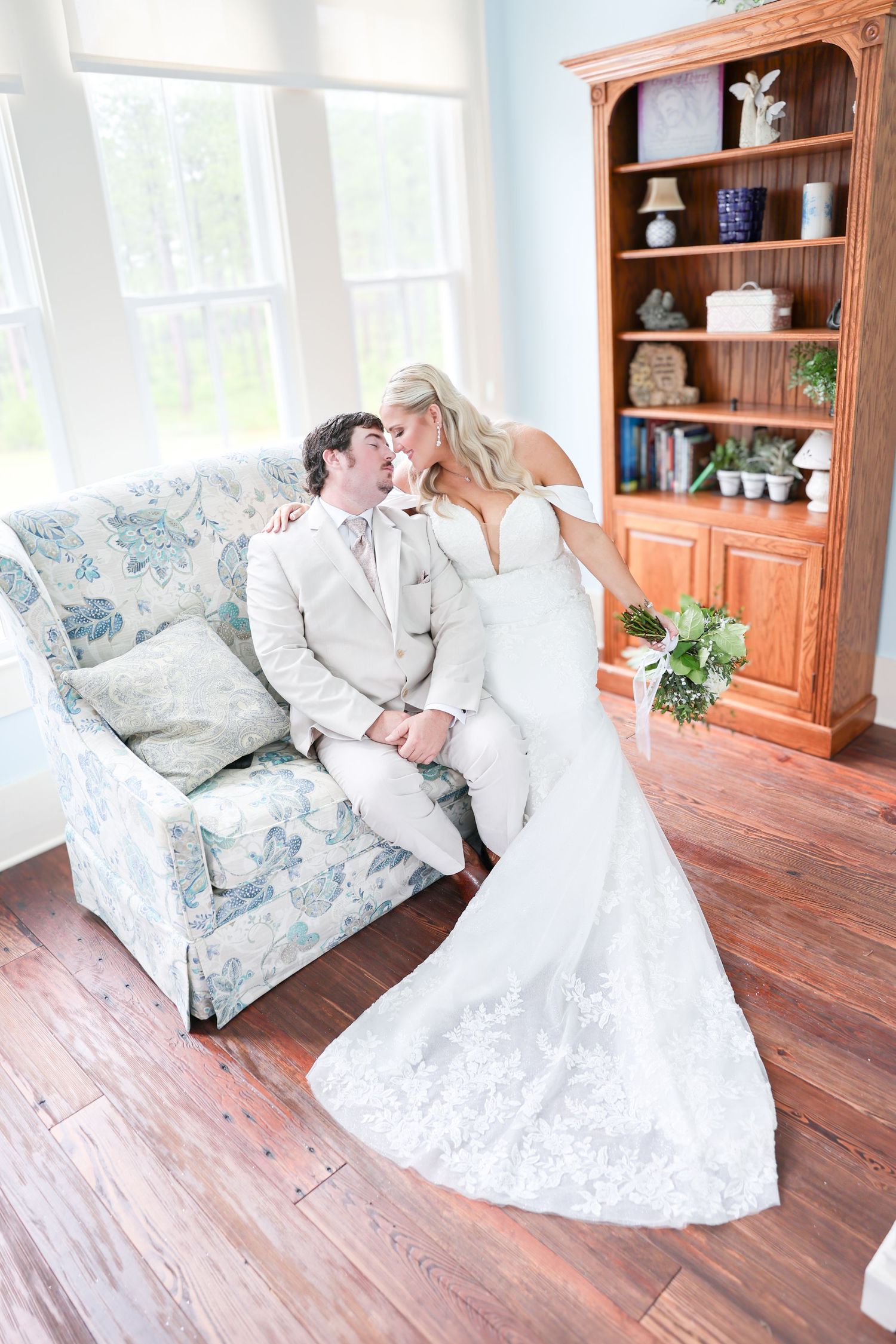 Bride in lace gown leans in to kiss groom in tan suit on floral sofa, holding bouquet, hardwood floors and bookshelf behind.