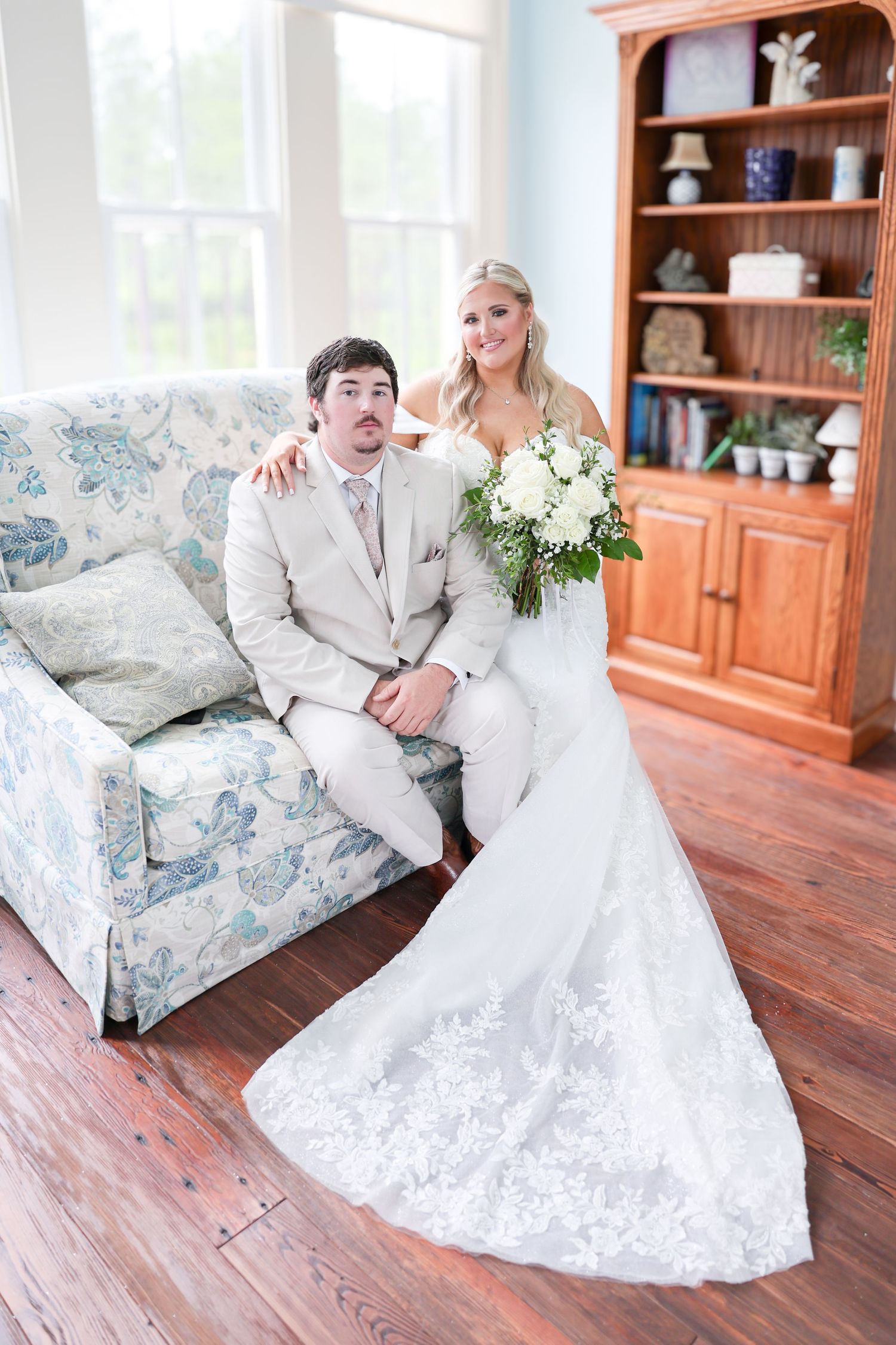 Bride in lace gown with bouquet posing with groom on floral sofa in rustic wood-floored room.