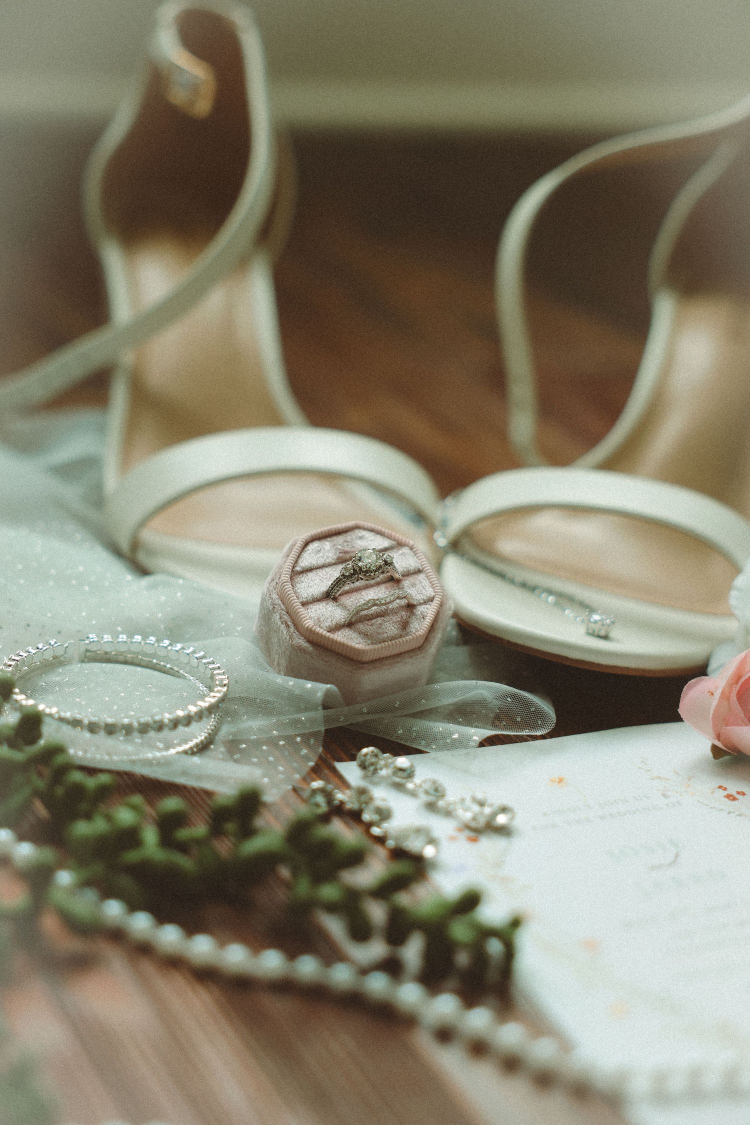 Elegant bridal flat lay featuring white heels, diamond ring in velvet box, pearl bracelet, earrings, and floral accents on wood.