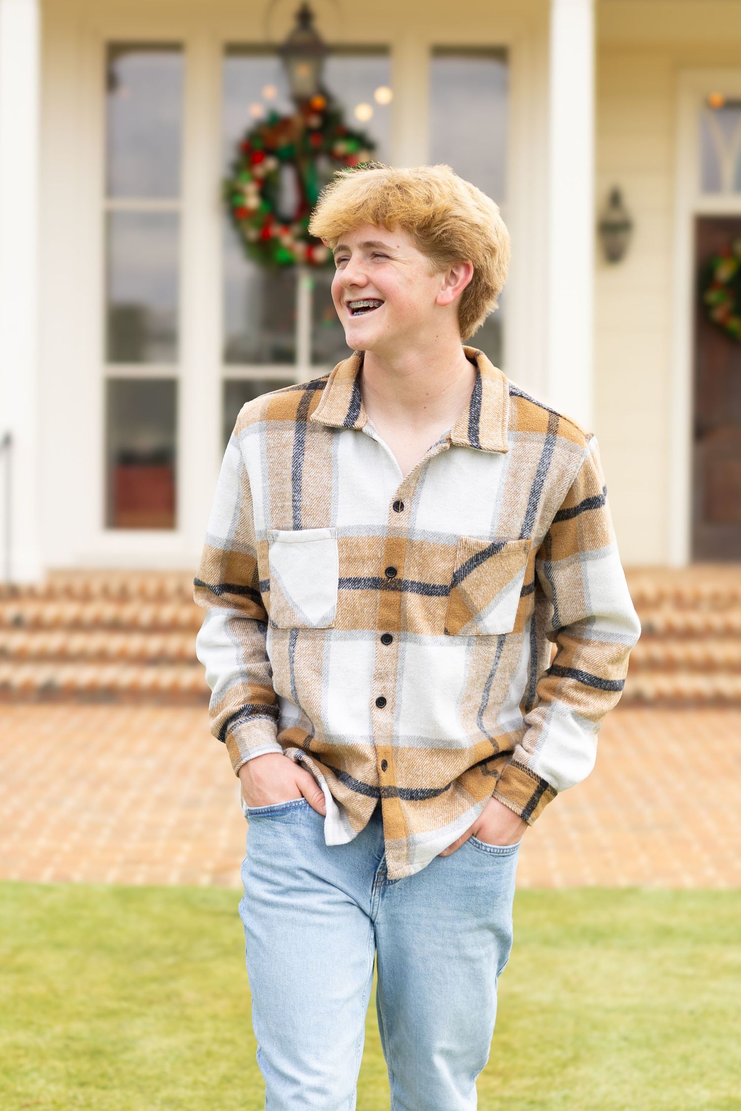 A person in a plaid shirt and jeans laughs joyfully while walking on grass in front of a brick house with holiday decor.