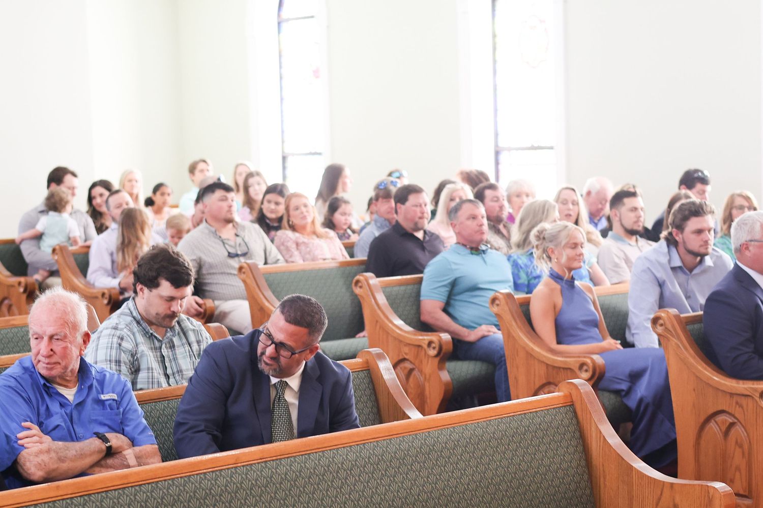 People sit in wooden pews during a church service or gathering in a bright, sunlit sanctuary.