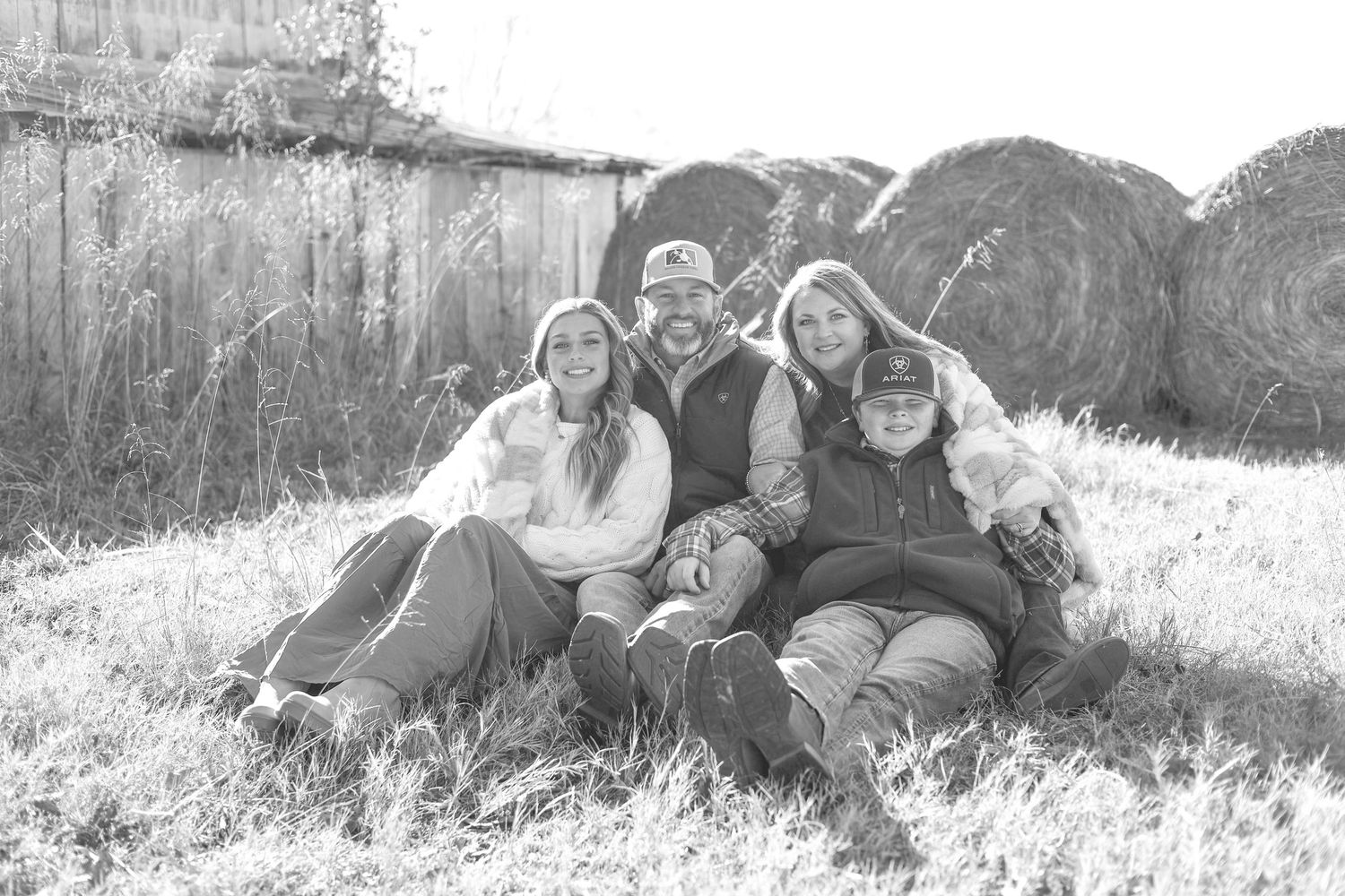 A black and white photo of a family group sitting together outdoors near hay bales on a sunny day.
