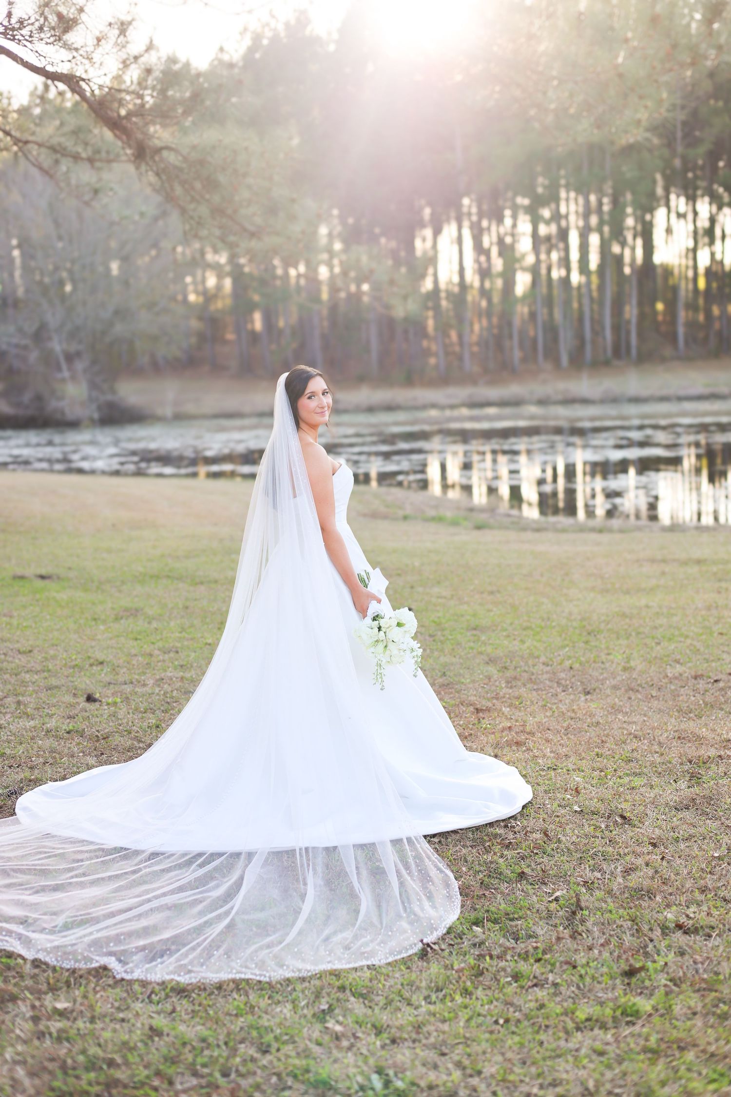 Bride in white gown with long cathedral veil poses near a sunlit pond surrounded by pine trees.