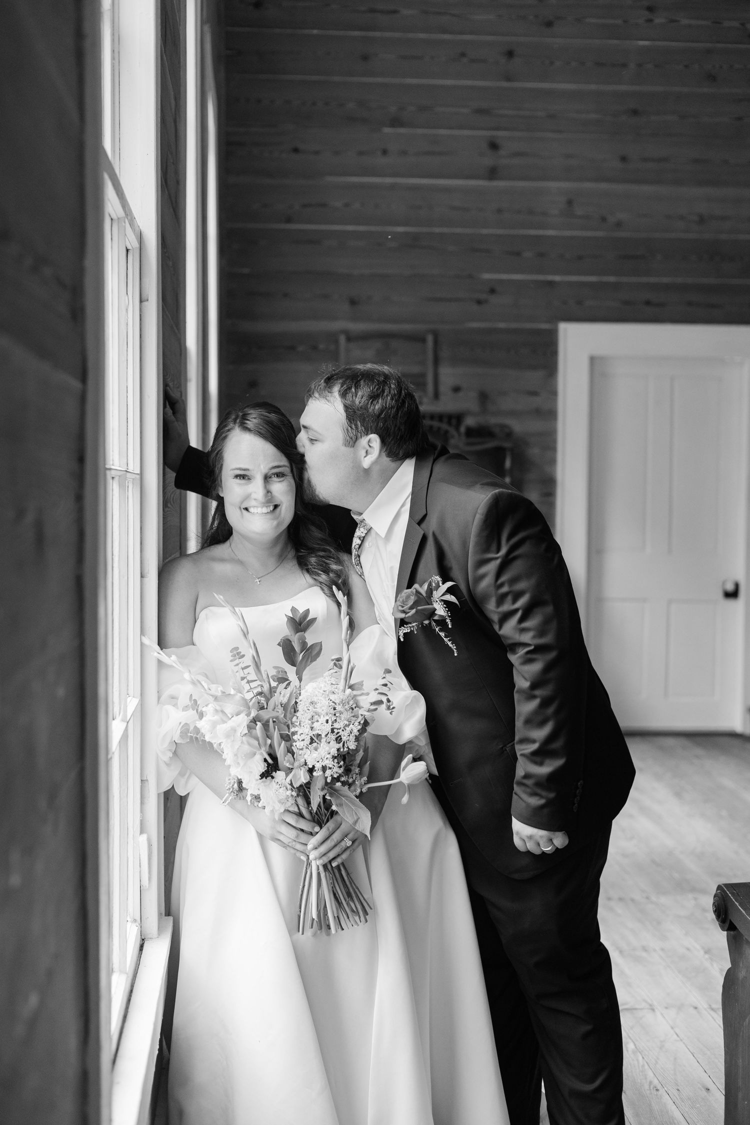 Groom in dark suit kissing bride on cheek as she smiles holding floral bouquet in black and white.