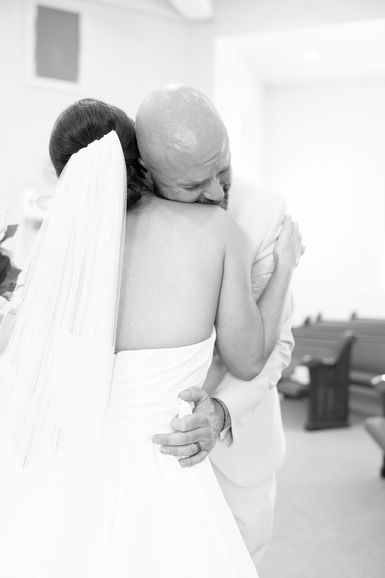 Emotional black and white photo of a bald father embracing his daughter in a wedding dress and veil inside a church.