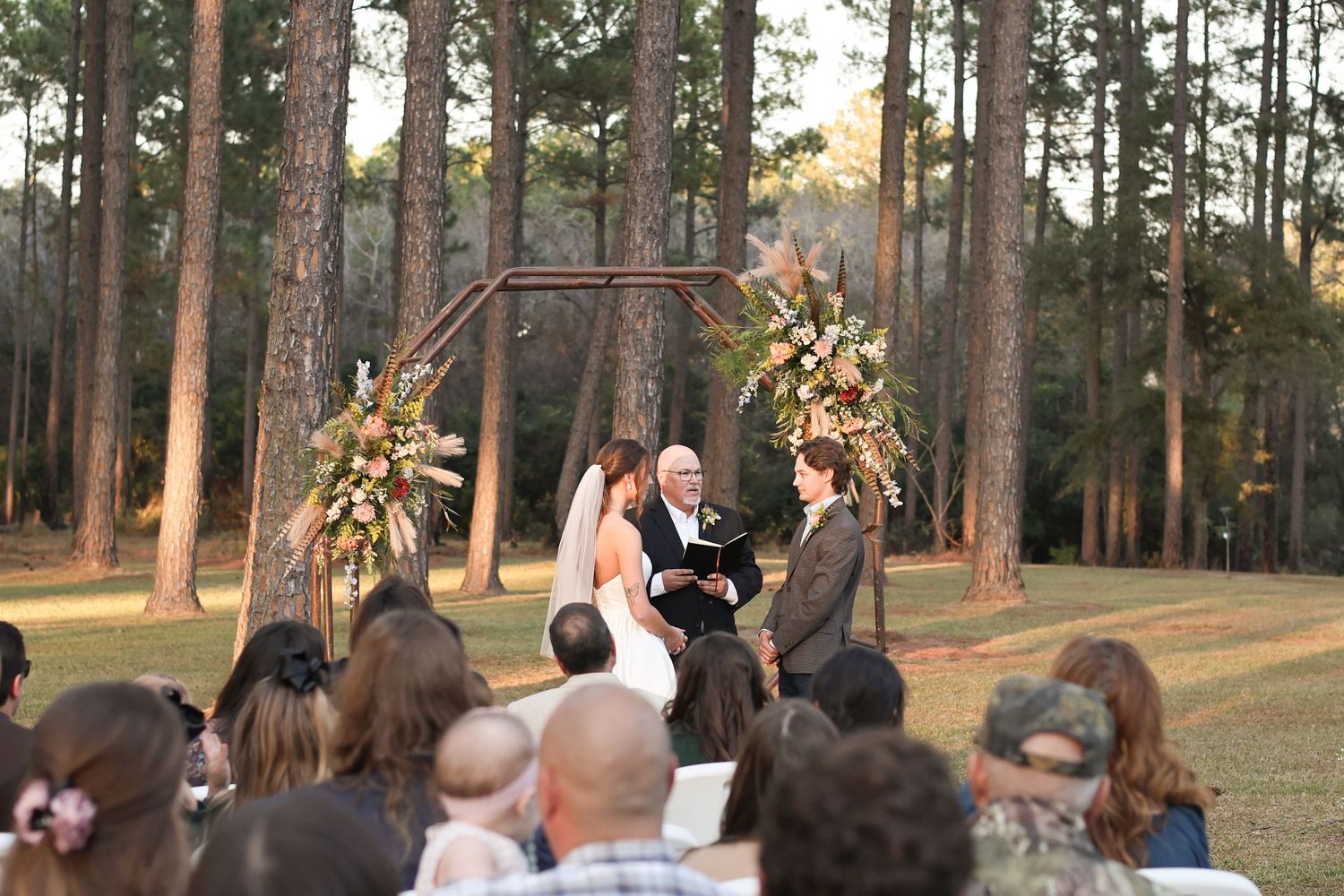 Wedding guests gather outdoors among tall pine trees for the ceremony.