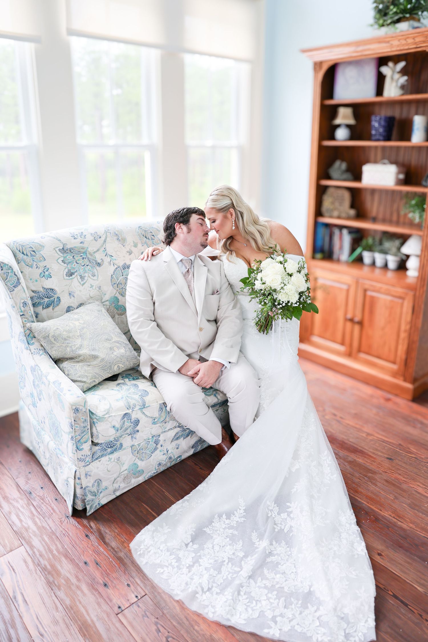 Bride and groom share a romantic moment on a floral sofa in a bright room with hardwood floors, bride holding white bouquet.