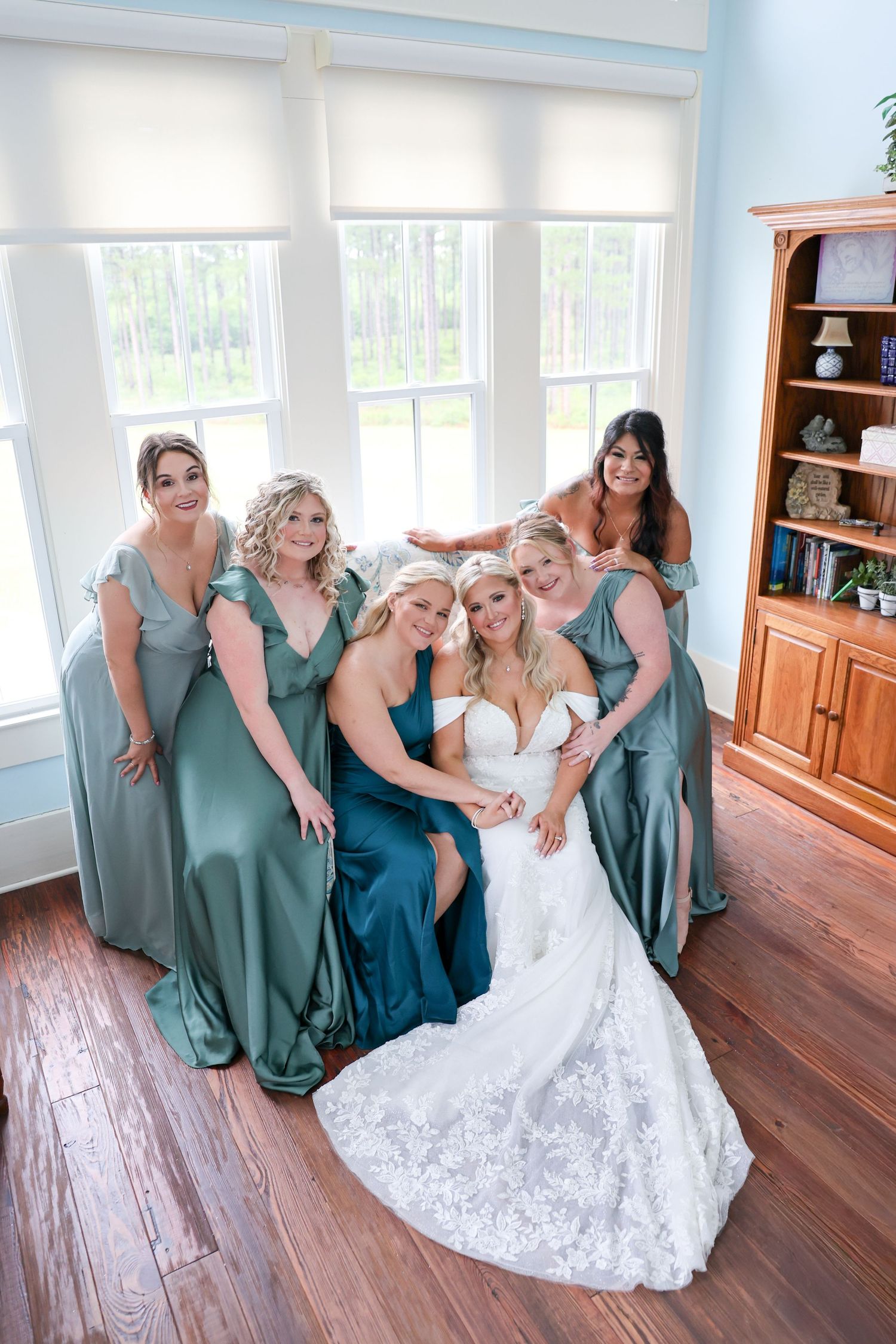 Bride in white lace gown poses with five bridesmaids in sage green dresses inside a bright room with hardwood floors.