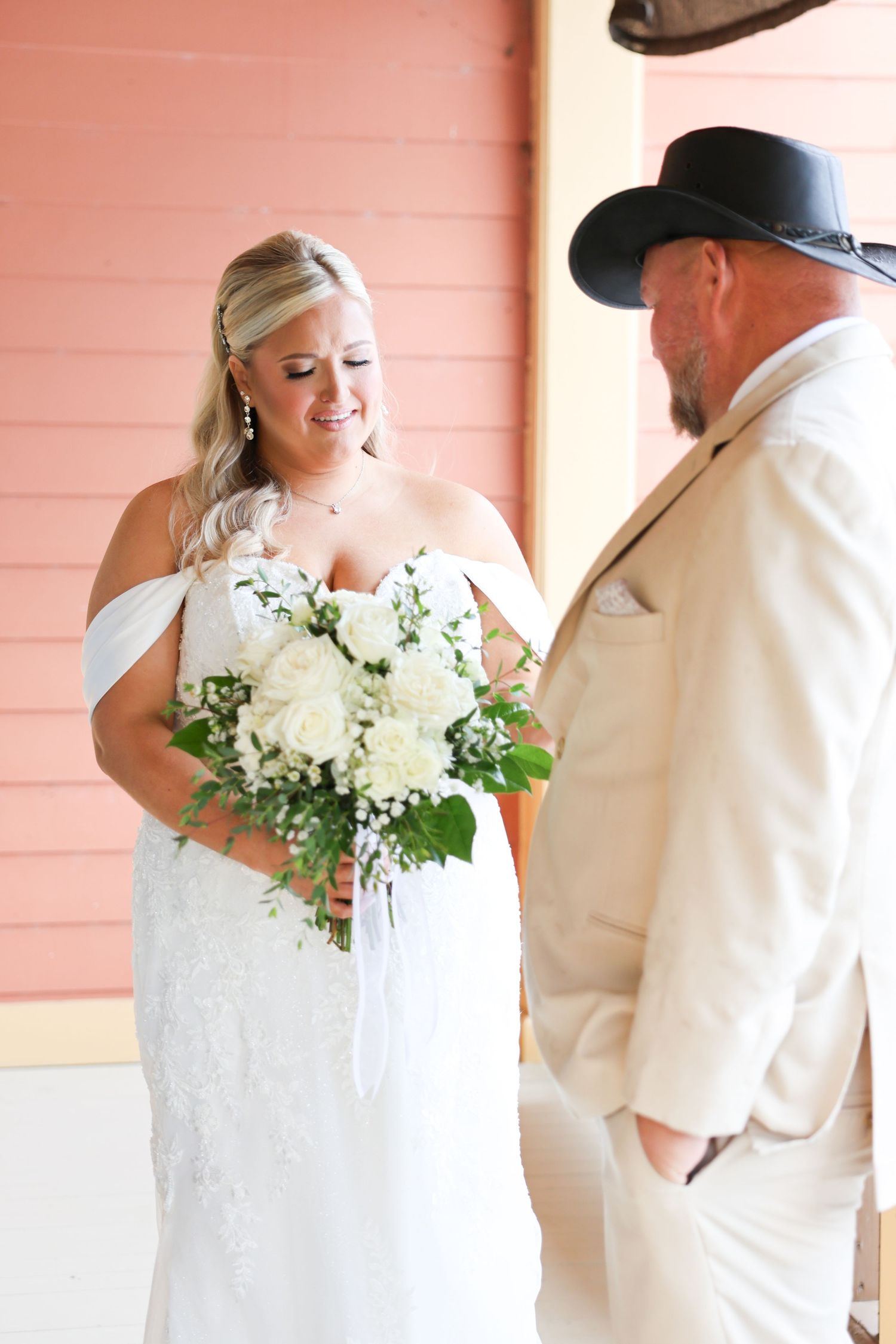 Bride in white off-shoulder lace gown holds white rose bouquet, smiling at groom in tan suit and cowboy hat.