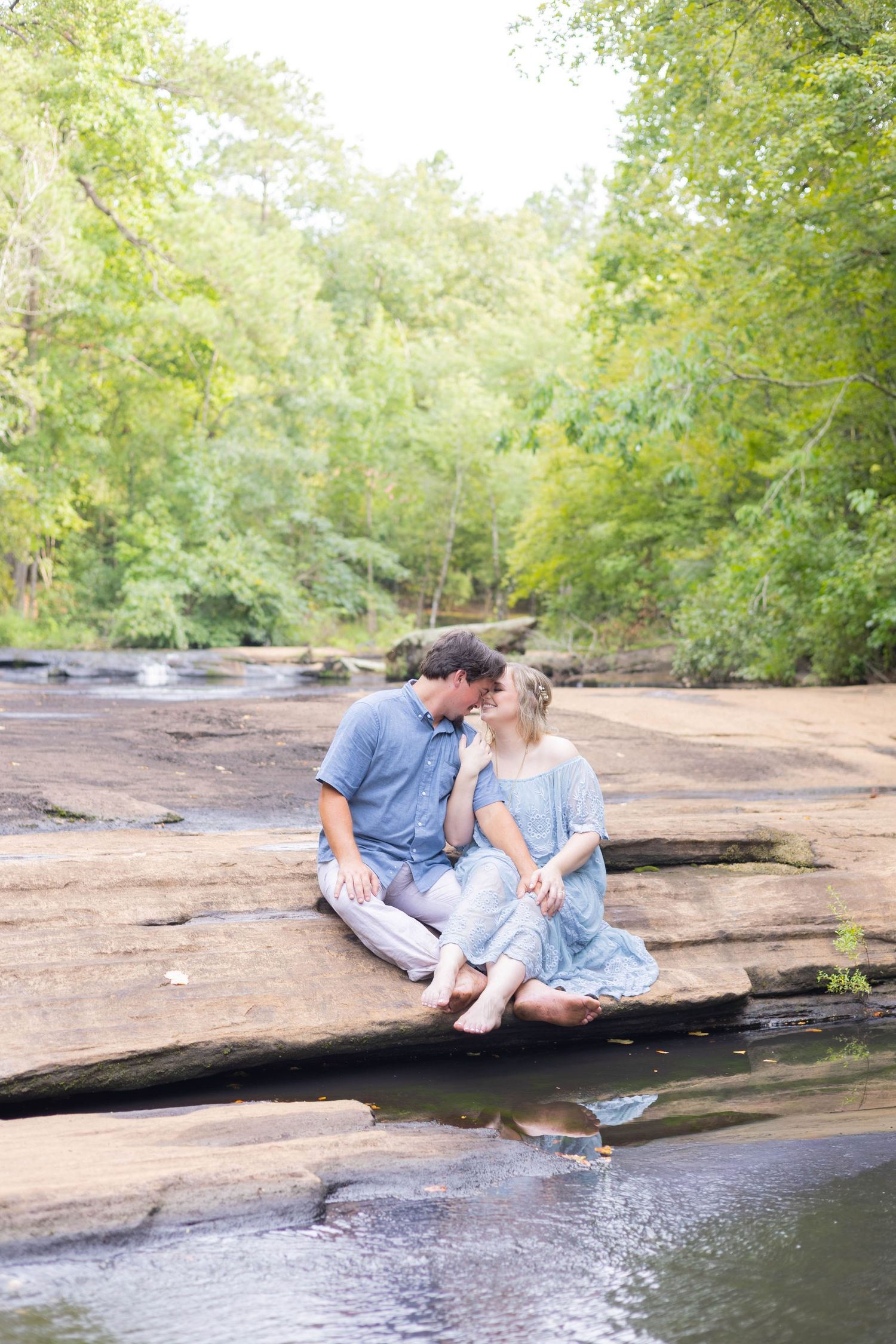 Couple sitting barefoot on rocky riverbank surrounded by lush green trees, sharing a romantic moment outdoors.