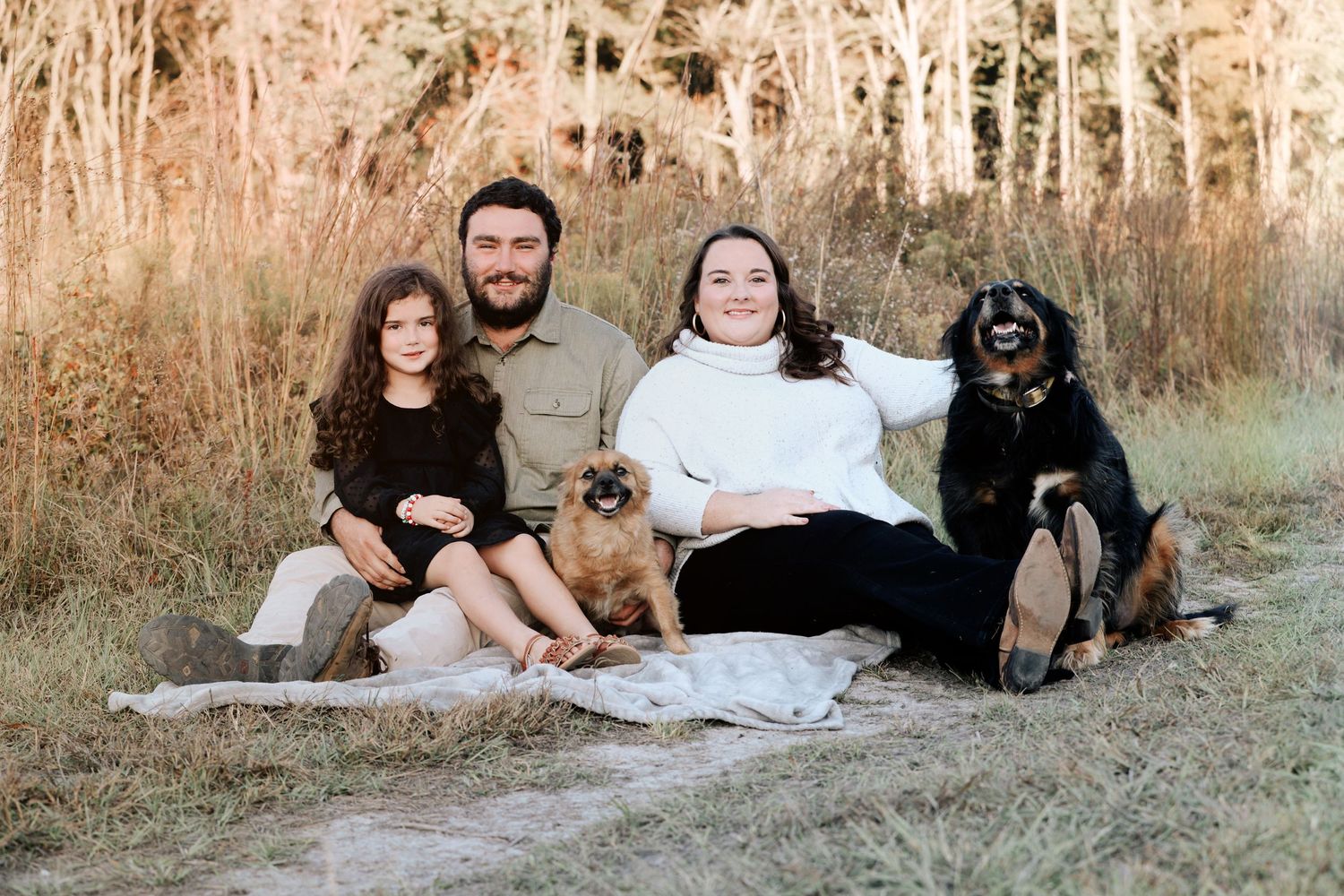 Family of two adults and a child sit outdoors on a blanket with two dogs, smiling in a natural wooded setting.