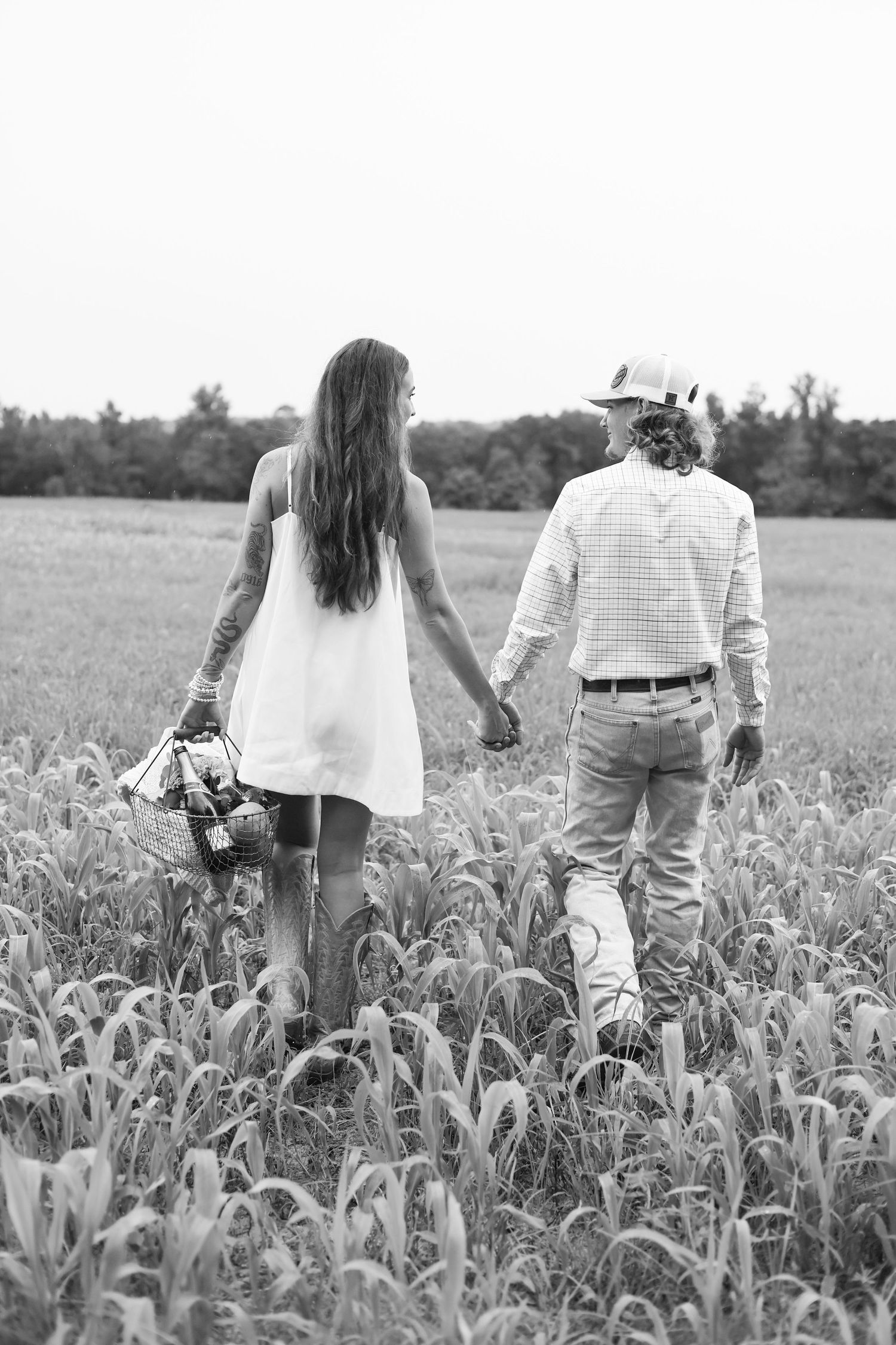 Black and white photo of a couple holding hands walking through a lush field, woman in white dress carrying flowers.