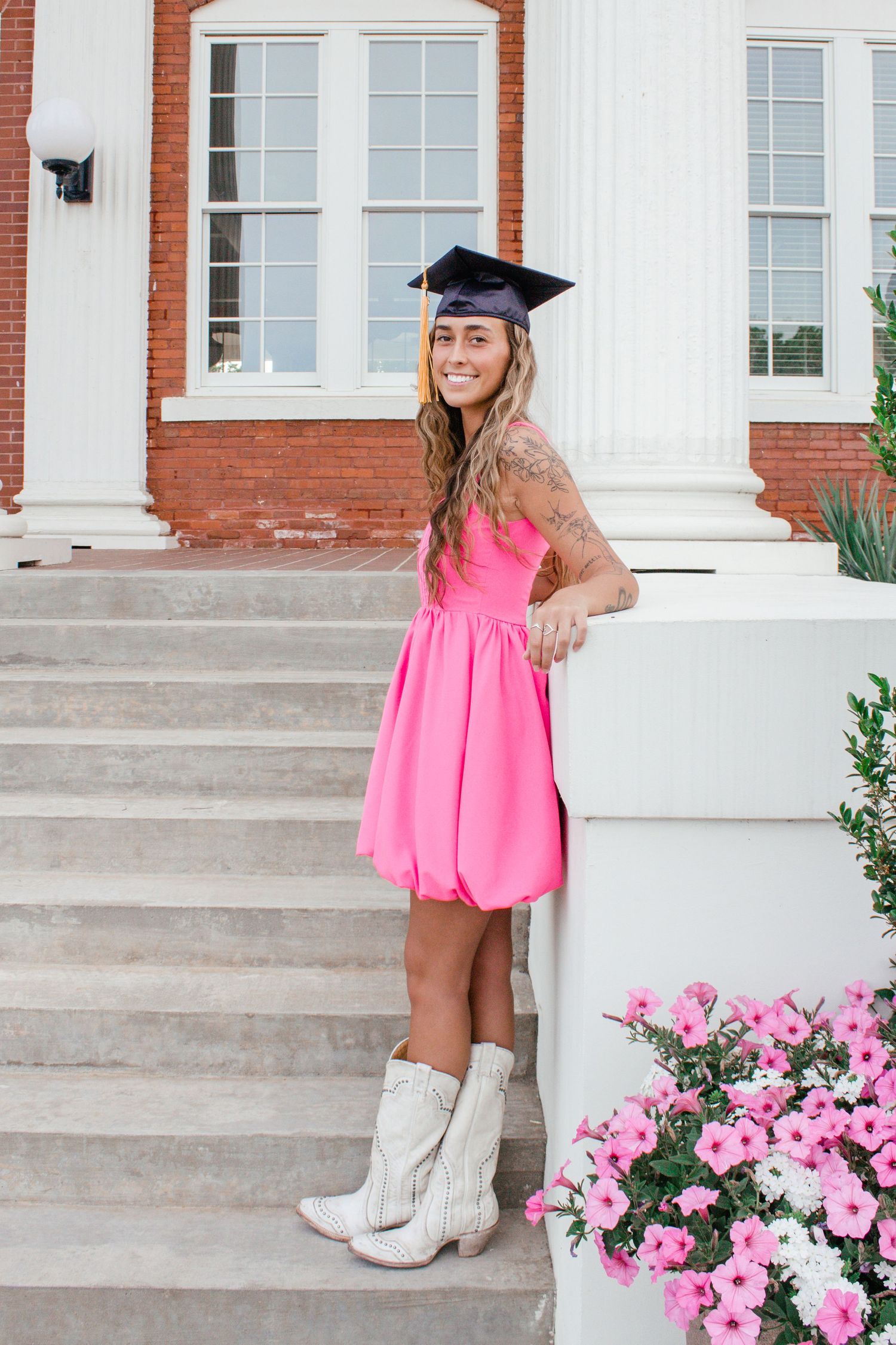 Smiling graduate in pink bubble dress and cowboy boots wearing cap on university steps with pink flowers.