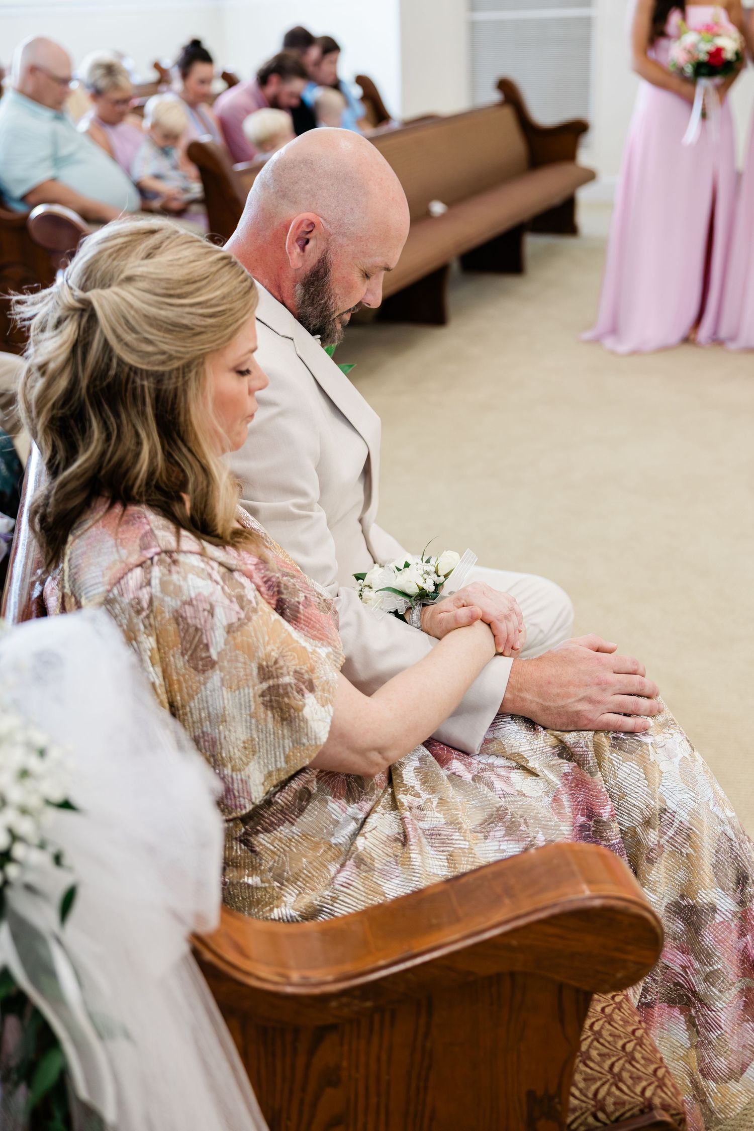 Couple in formal attire sit together on church pew during wedding ceremony, woman in floral dress holding small bouquet.