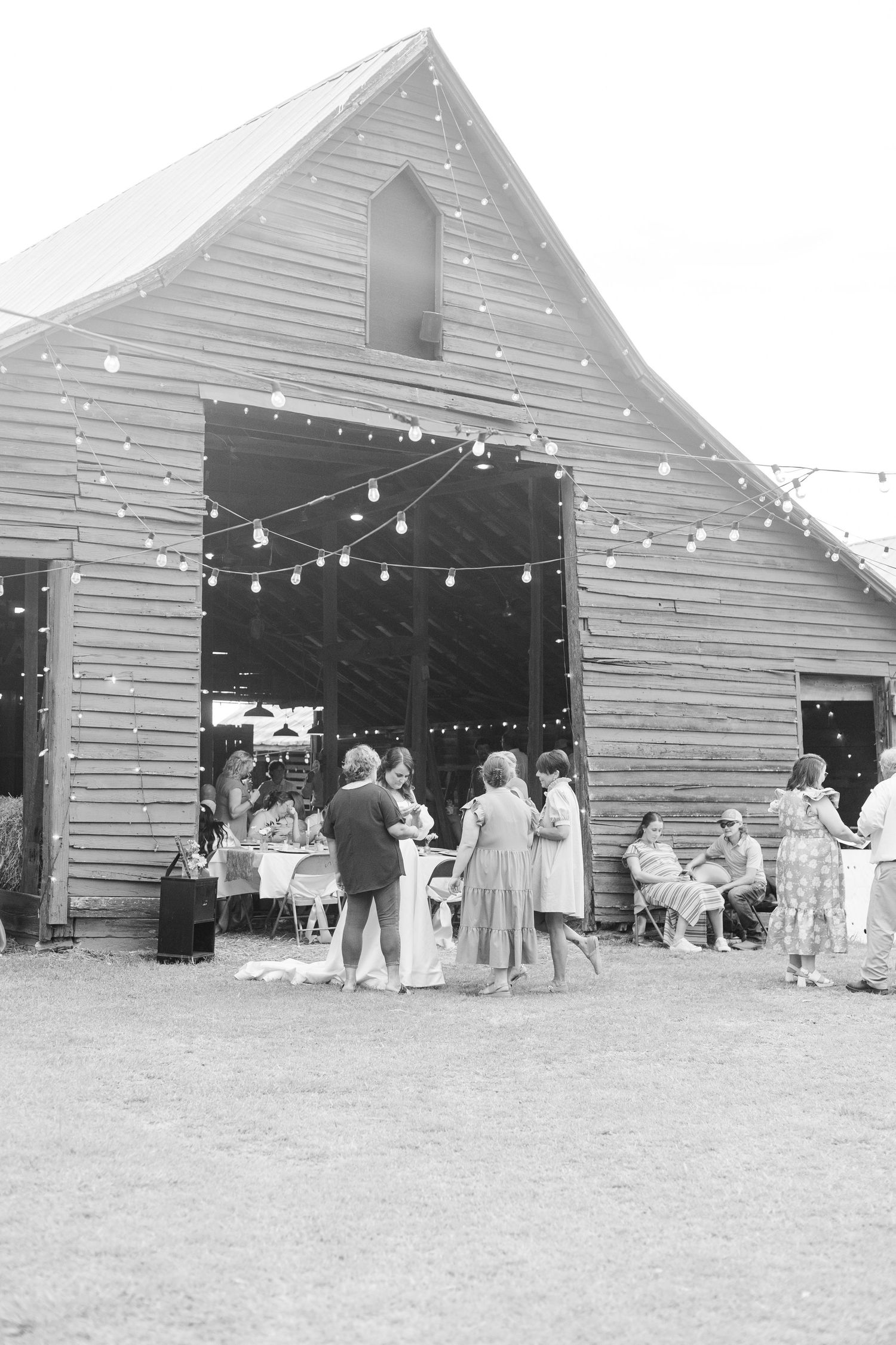 Wedding guests mingle outside rustic wooden barn decorated with string lights at outdoor reception in black and white.