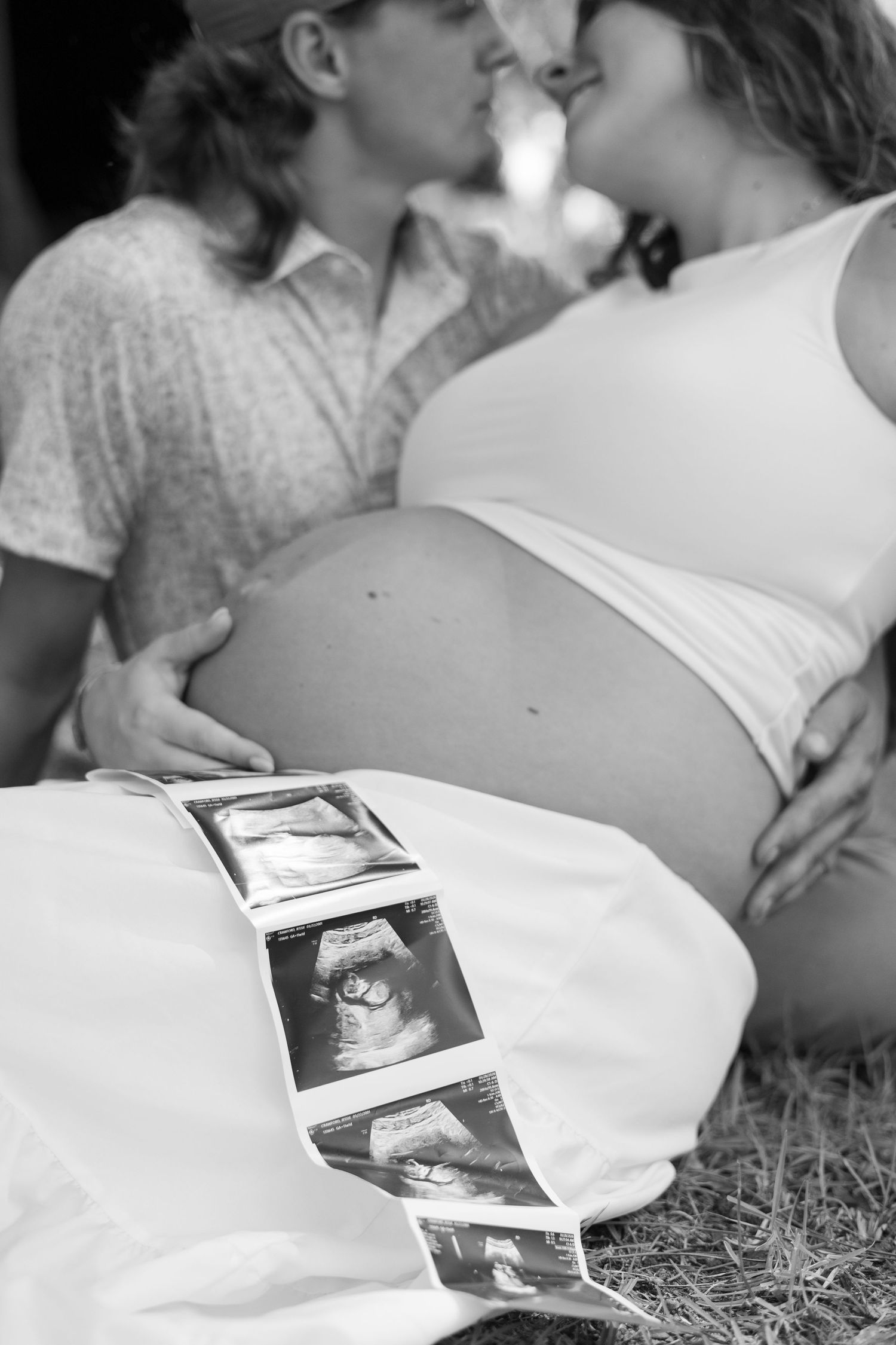 Black and white photo of pregnant couple sharing a kiss with ultrasound photos resting on baby bump.