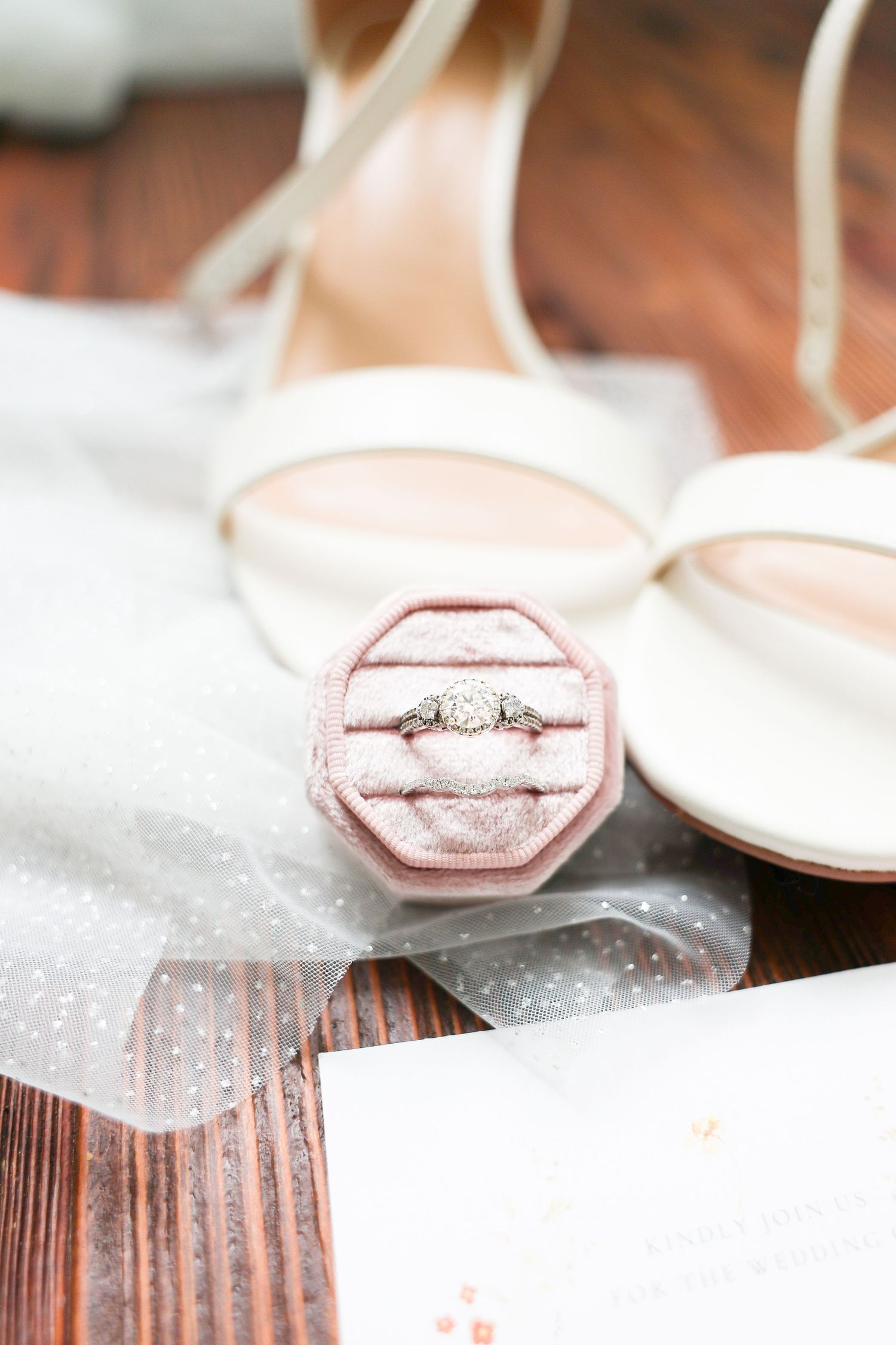Diamond engagement ring in pink velvet octagonal box beside white strappy heels on wooden floor.