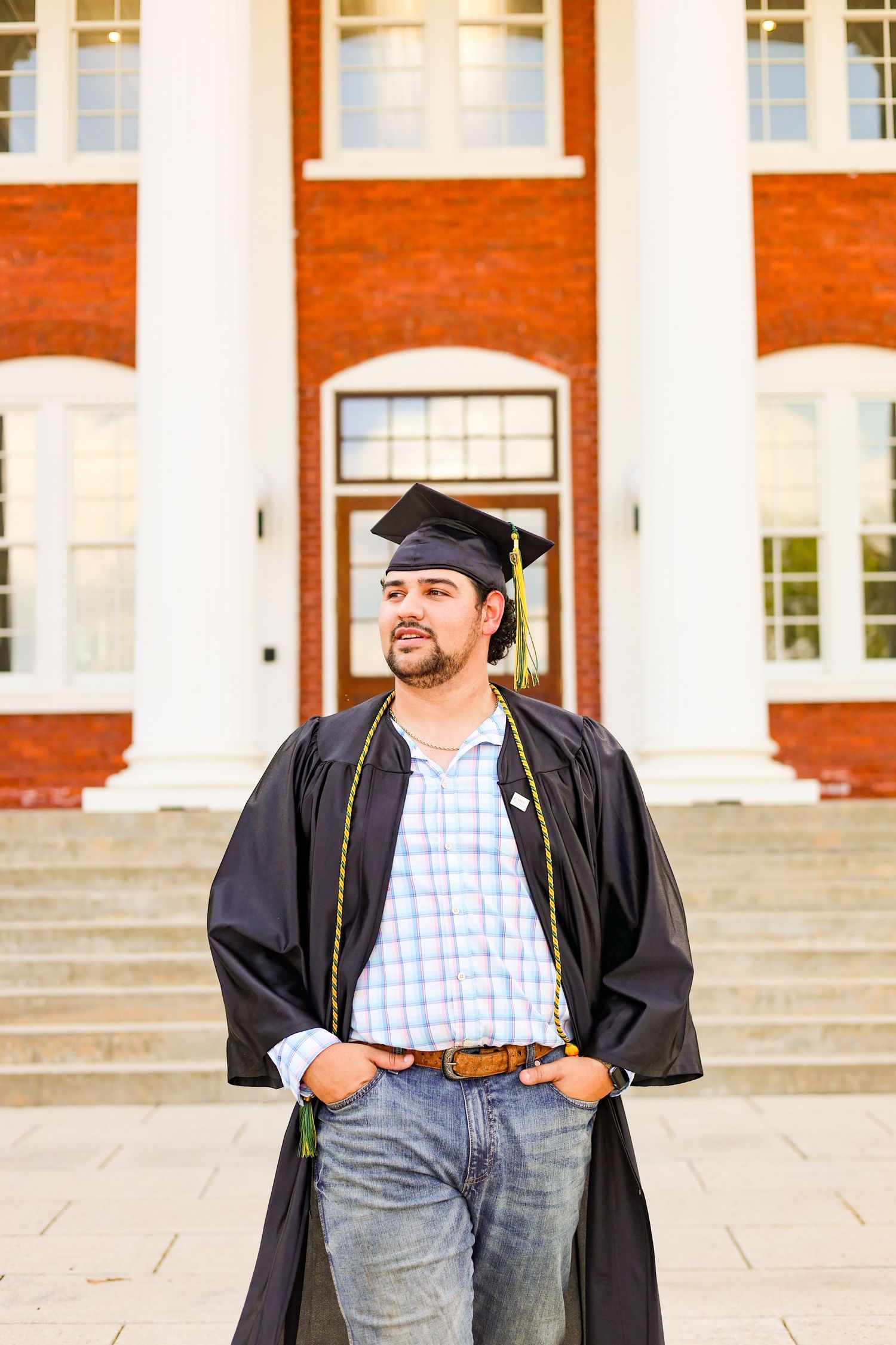 Graduate in cap and gown poses in front of red brick building with white columns.