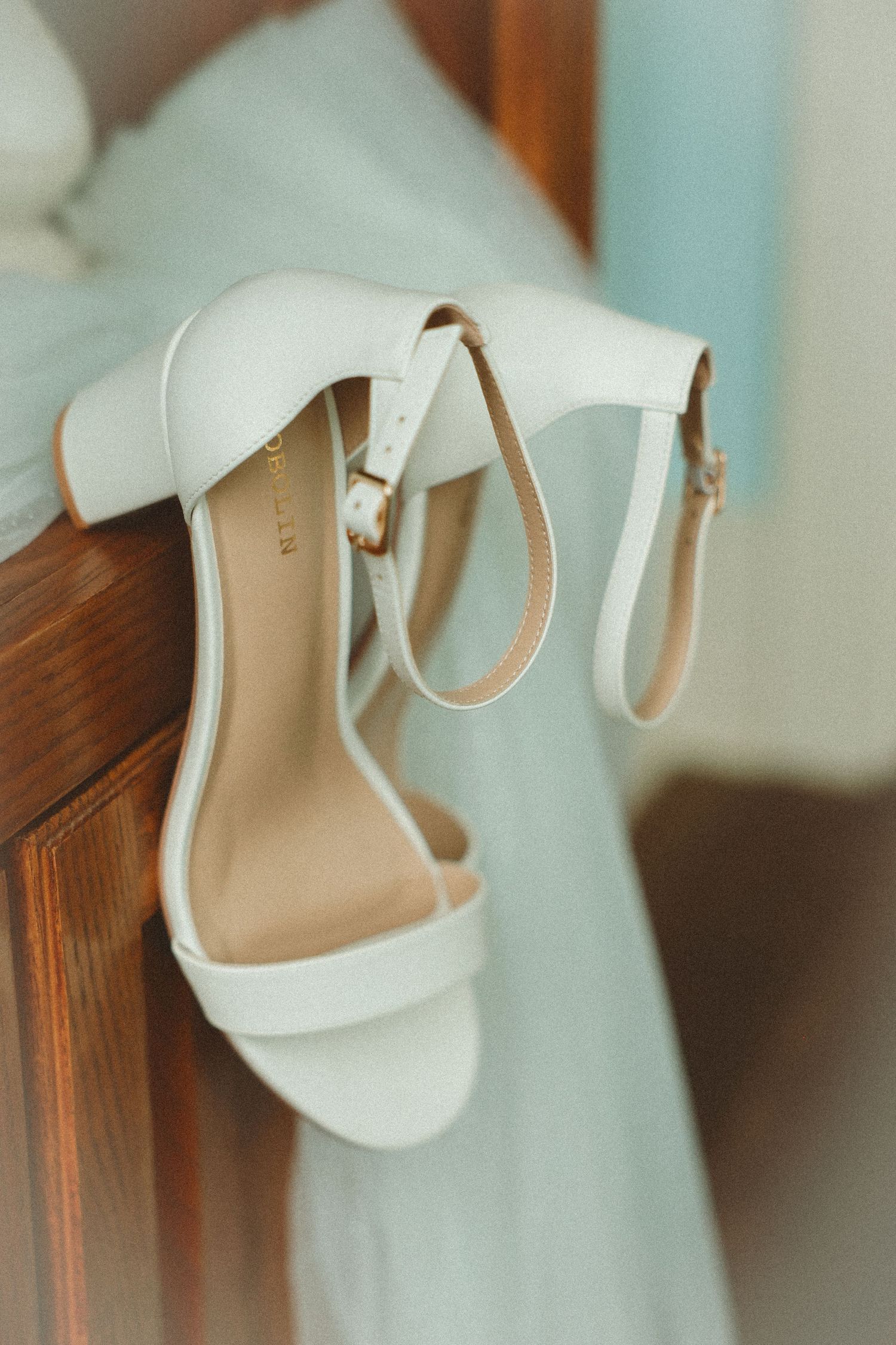 Elegant white bridal heeled sandals with ankle strap resting on wooden chair with soft blue dress.