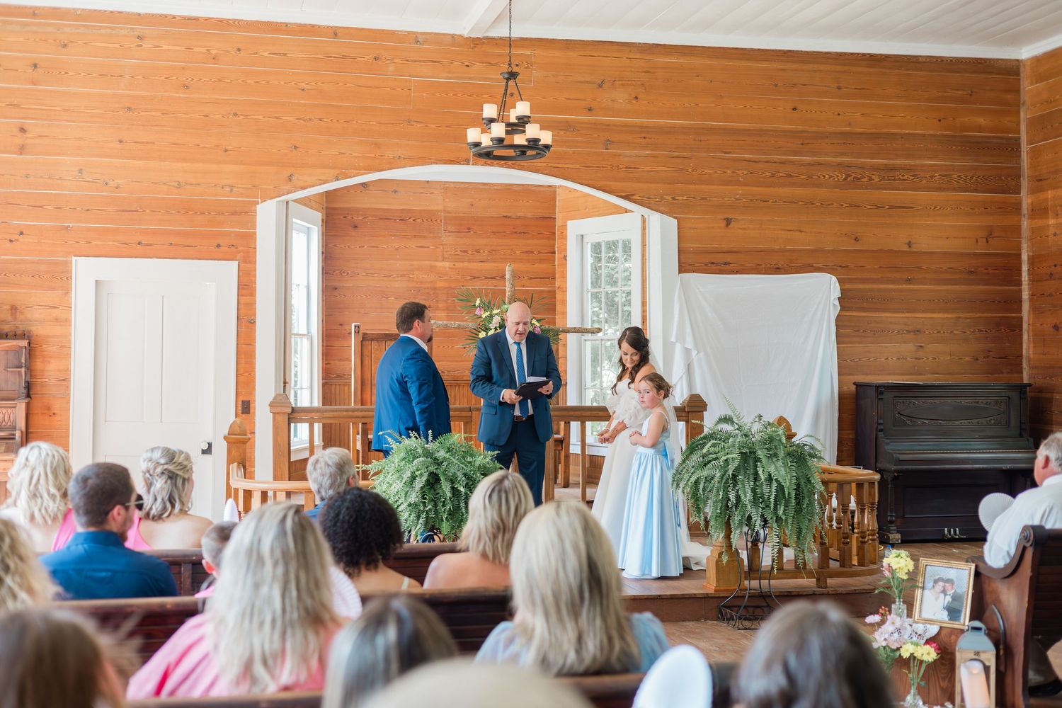 Bride and groom exchange vows at rustic wood-paneled chapel altar with officiant, guests seated in pews.