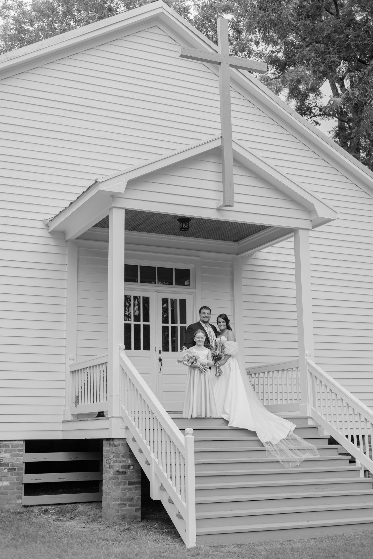 Bride and groom pose with officiant on steps of a charming white chapel with a cross at their wedding.