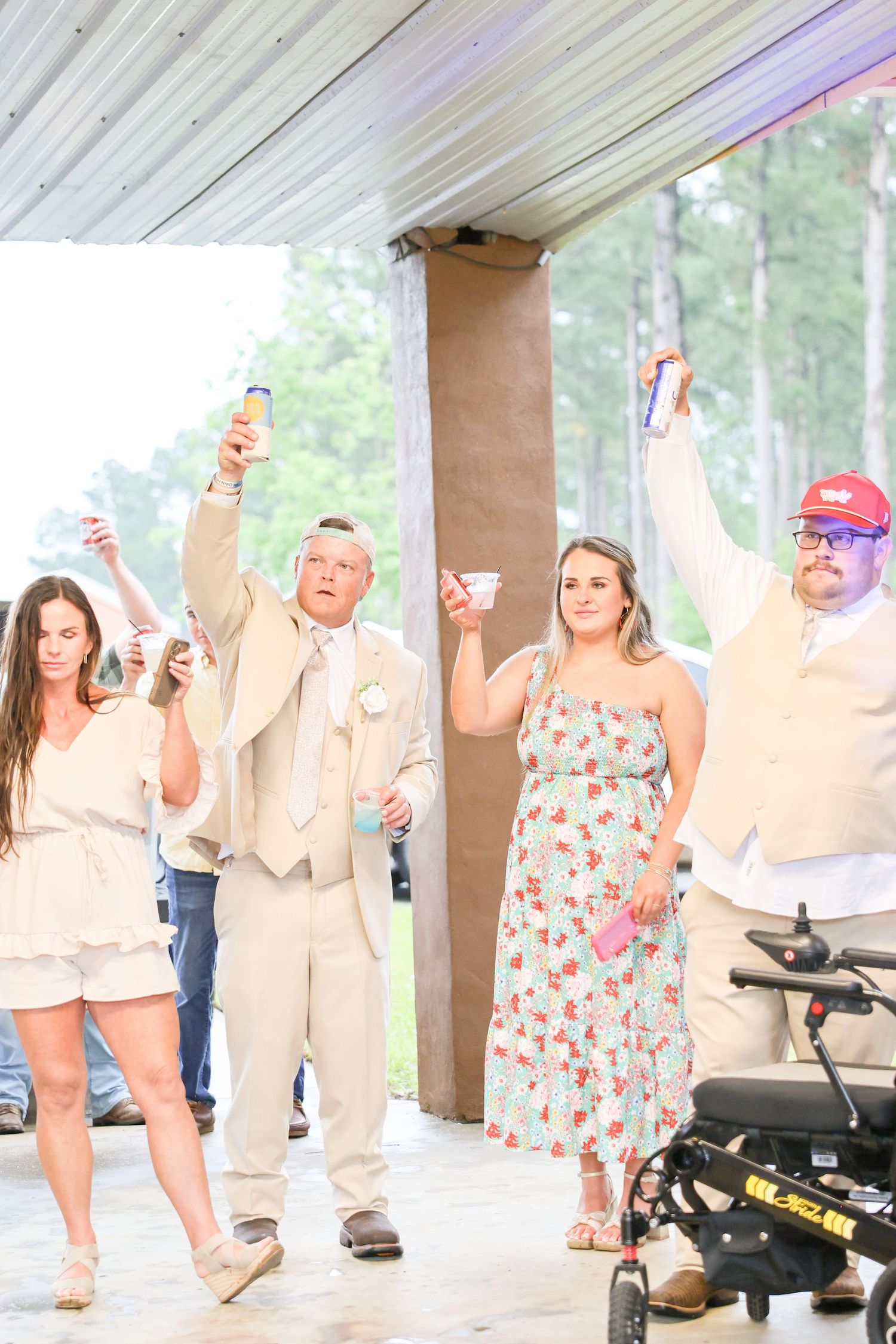 Wedding guests raising drinks in a toast outdoors under a pavilion surrounded by trees.