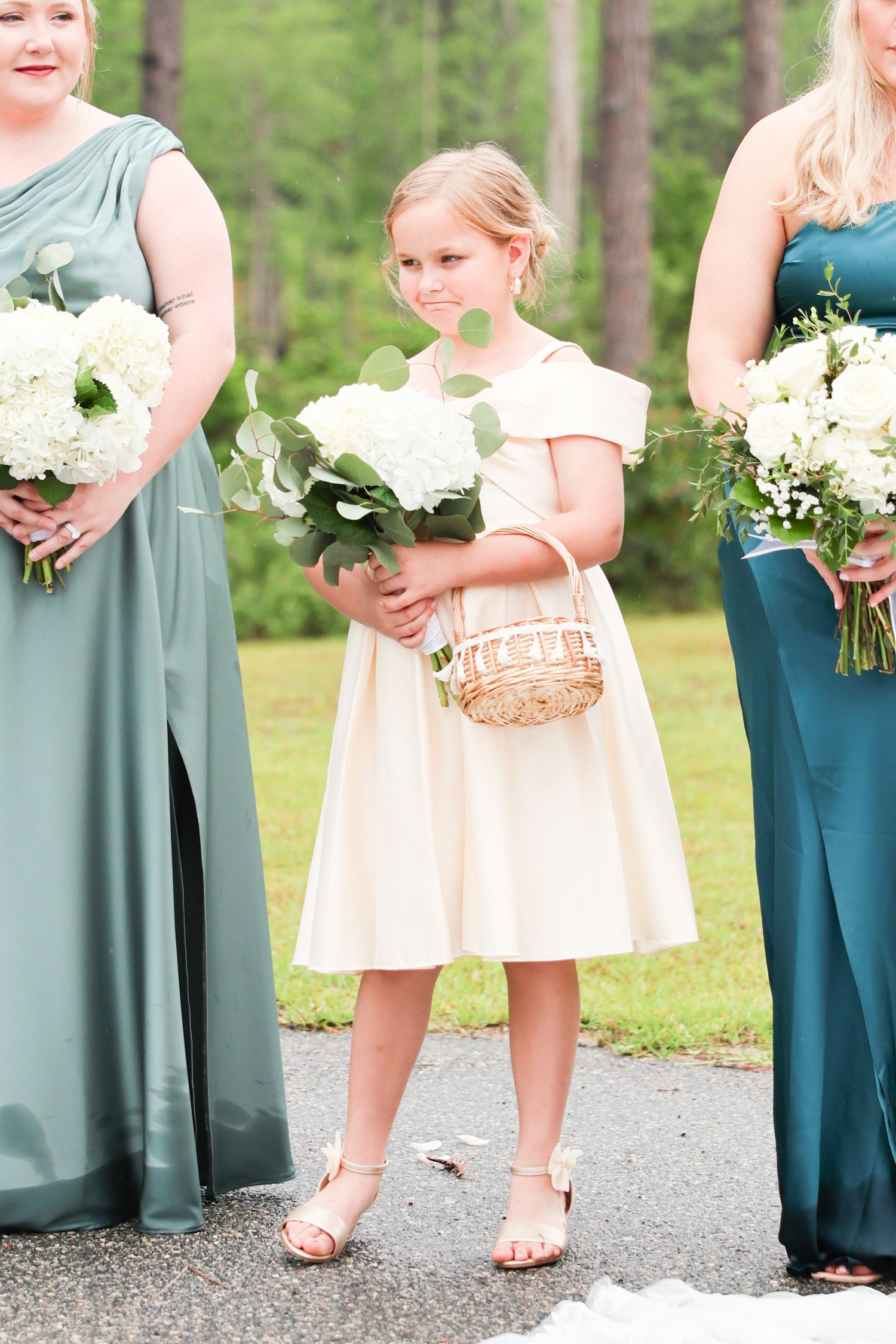 Young flower girl in cream dress holds white hydrangeas and wicker basket, flanked by bridesmaids in green gowns.