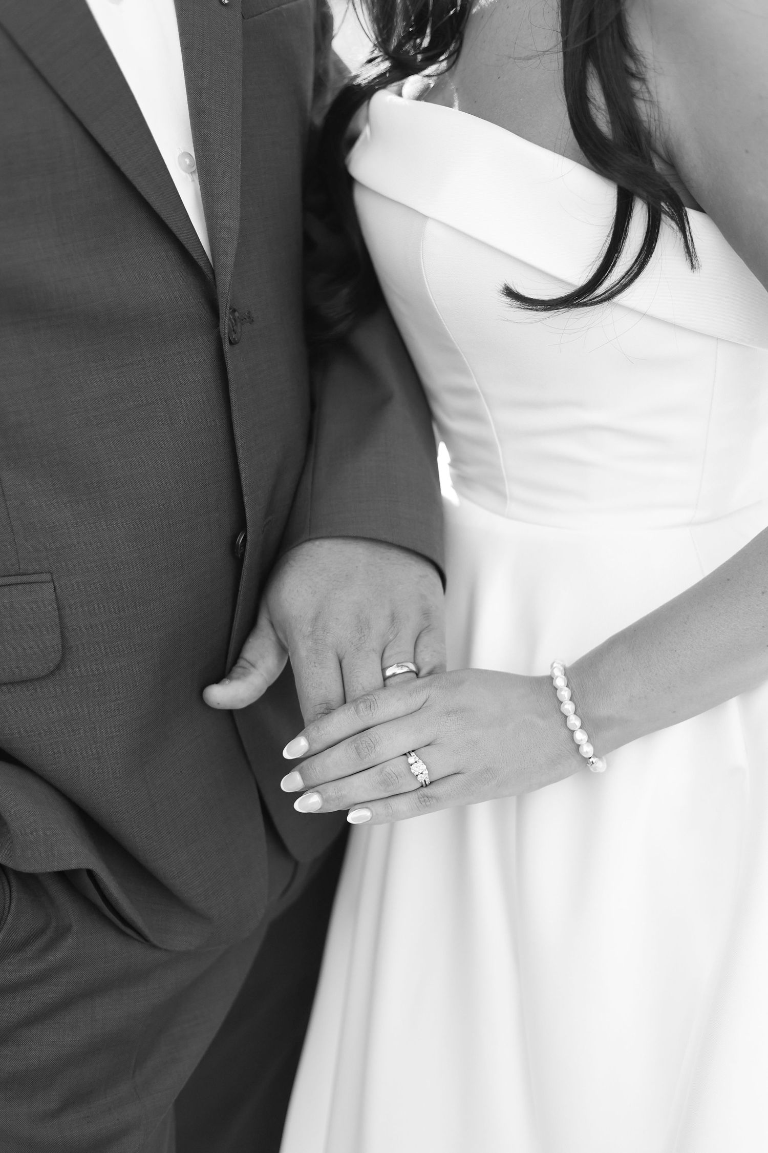 Newlyweds display their wedding rings while holding hands, bride wearing pearl bracelet and white gown.