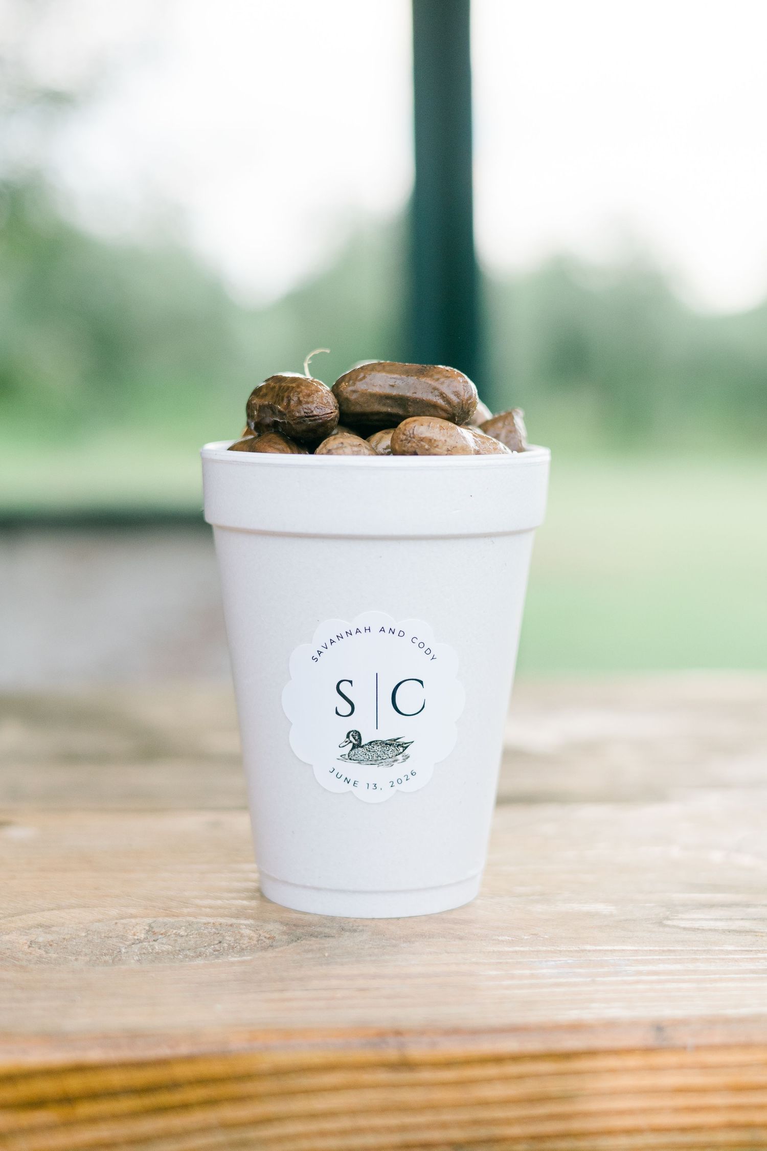 White foam cup with S|C monogram and duck logo filled with stones, sitting on wooden table at outdoor wedding.