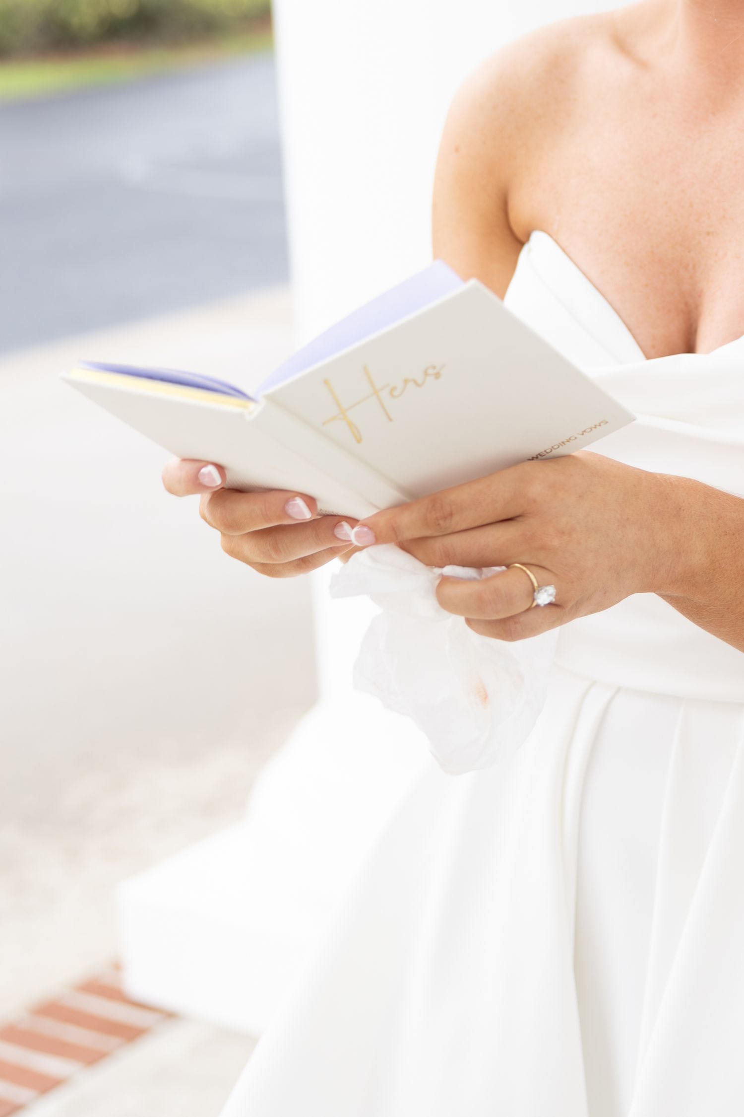 Bride in white dress reading wedding vows from a small book with gold script, wearing an engagement ring.