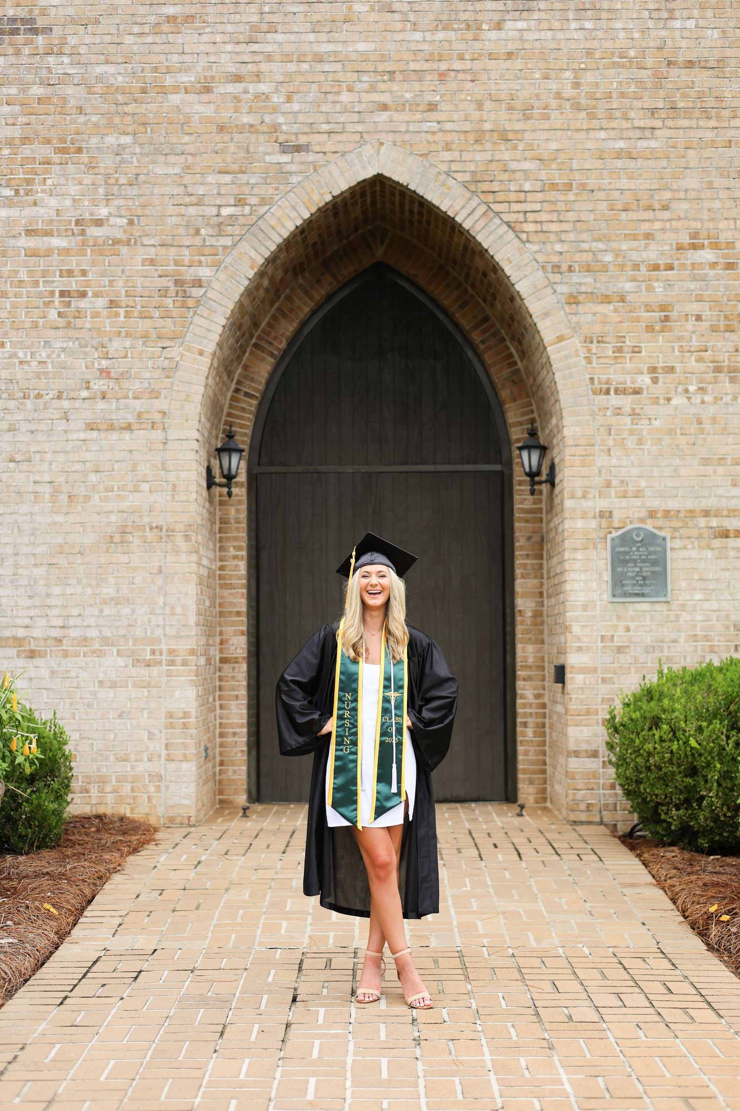 Graduate in cap and gown poses under arched brick doorway.