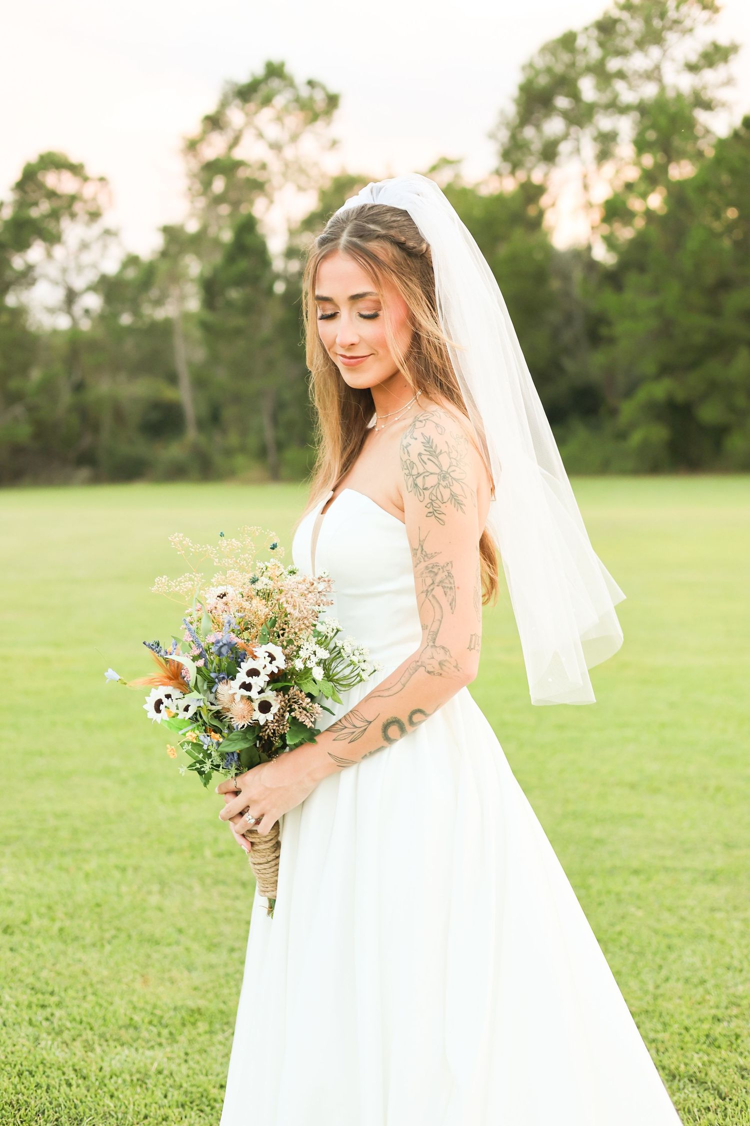 A bride poses with a wildflower bouquet against a backdrop of trees during sunset.