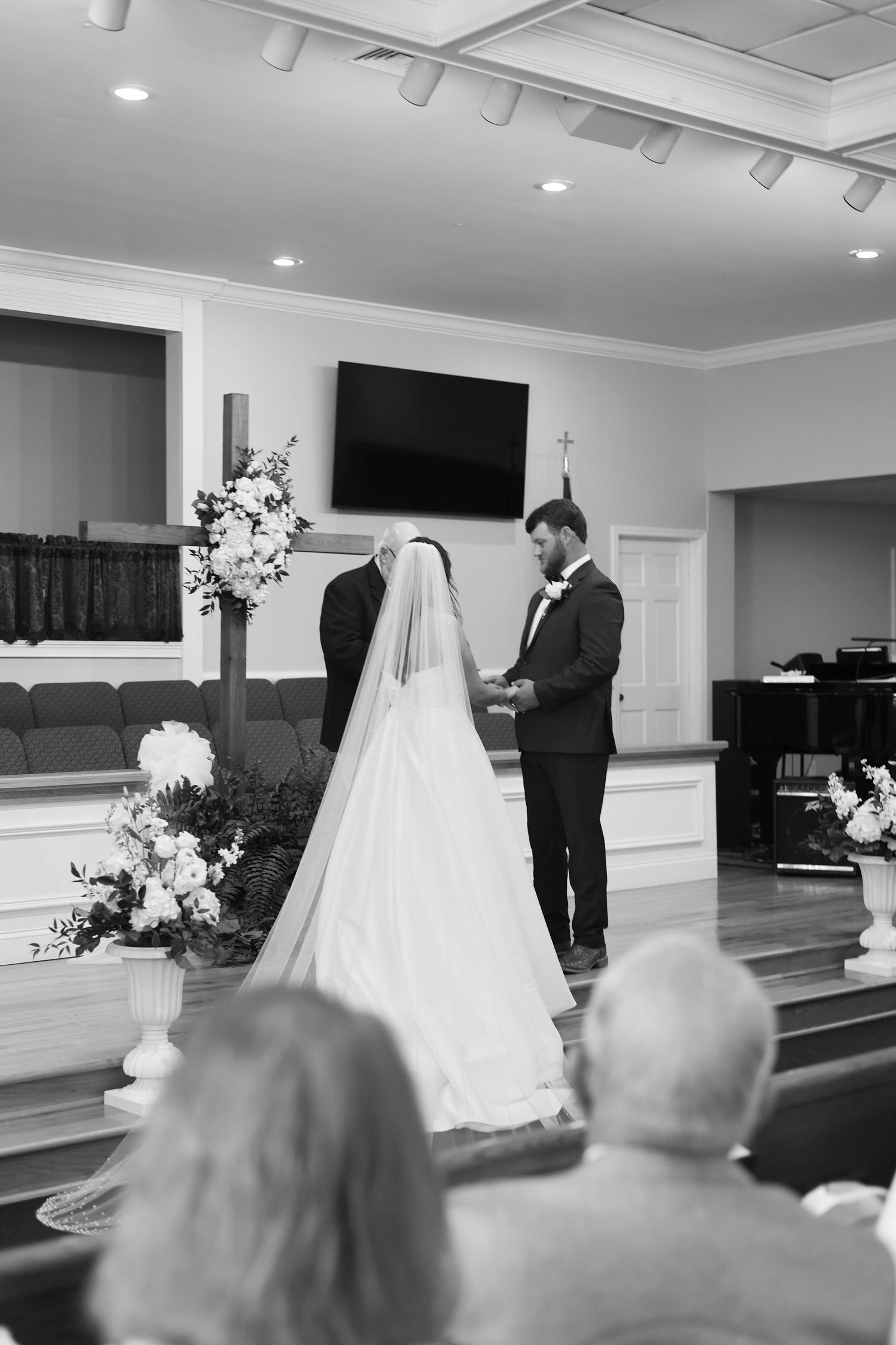 Black and white photo of a bride and groom exchanging vows at a church altar adorned with floral arrangements.