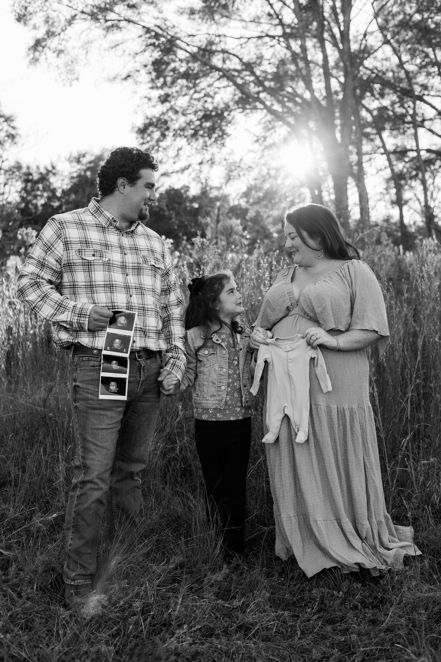 Black and white family maternity photo with parents and child holding ultrasound photos outdoors in a sunlit field.