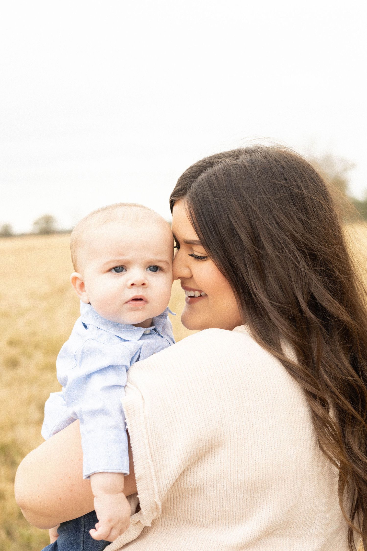 A young brunette mother tenderly nuzzles her smiling baby boy in a sunlit golden field.