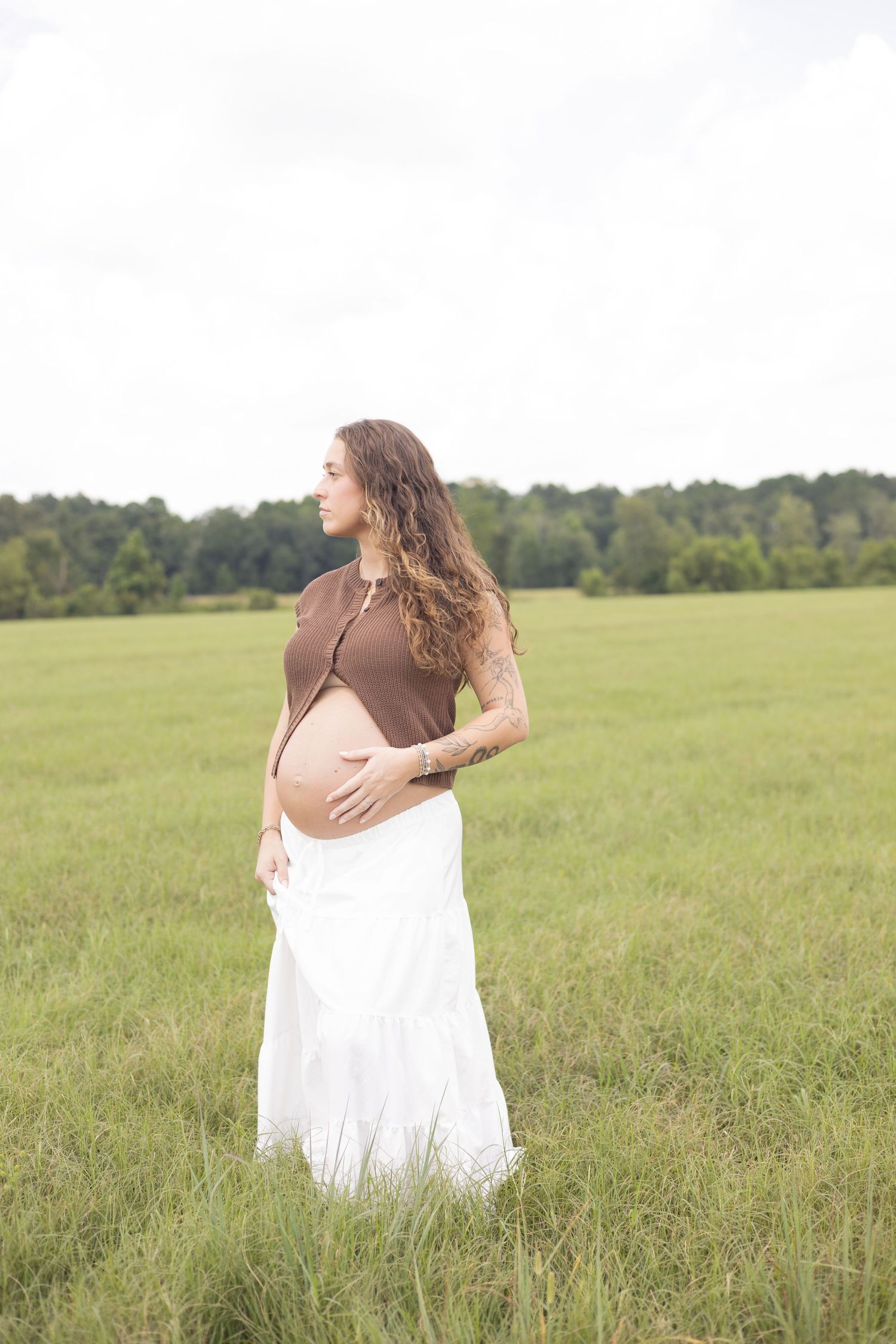 Pregnant woman in brown crop top and white skirt cradling baby bump in a lush green field.