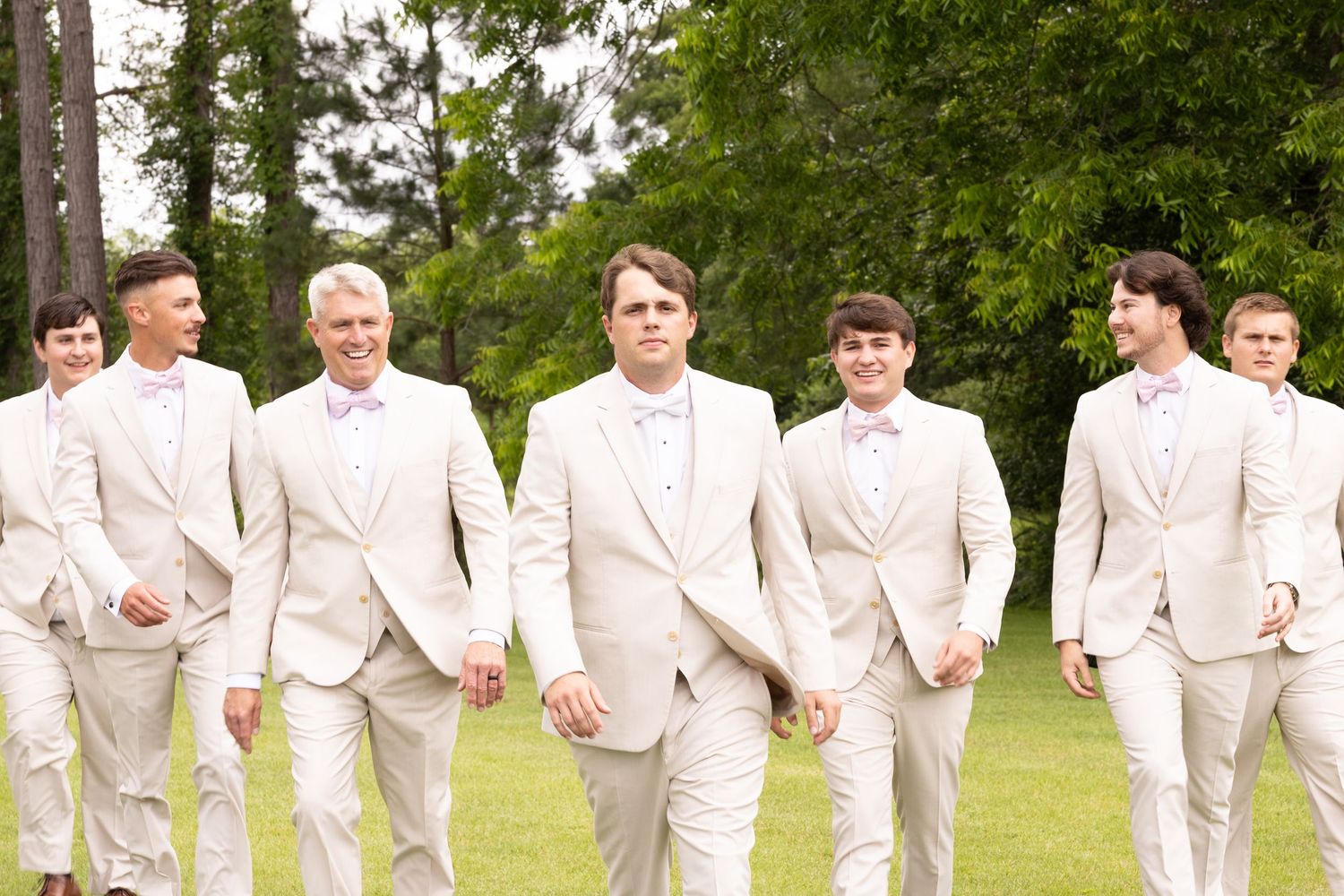 Groom and groomsmen in matching ivory suits with pink bow ties walking outdoors on green grass at a wedding.