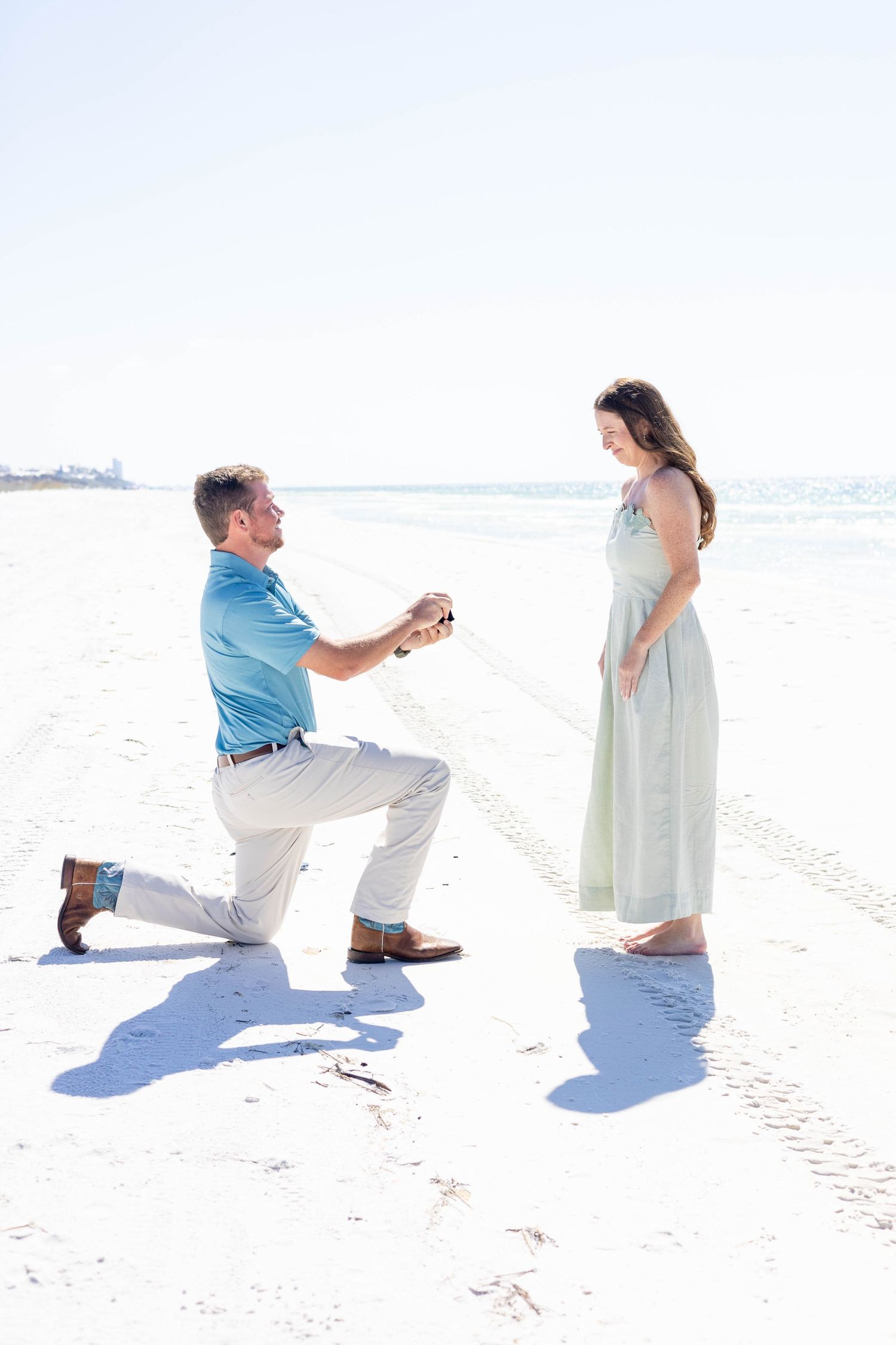 Man proposing on one knee to woman on a white sandy beach, holding a ring box in a romantic setting.