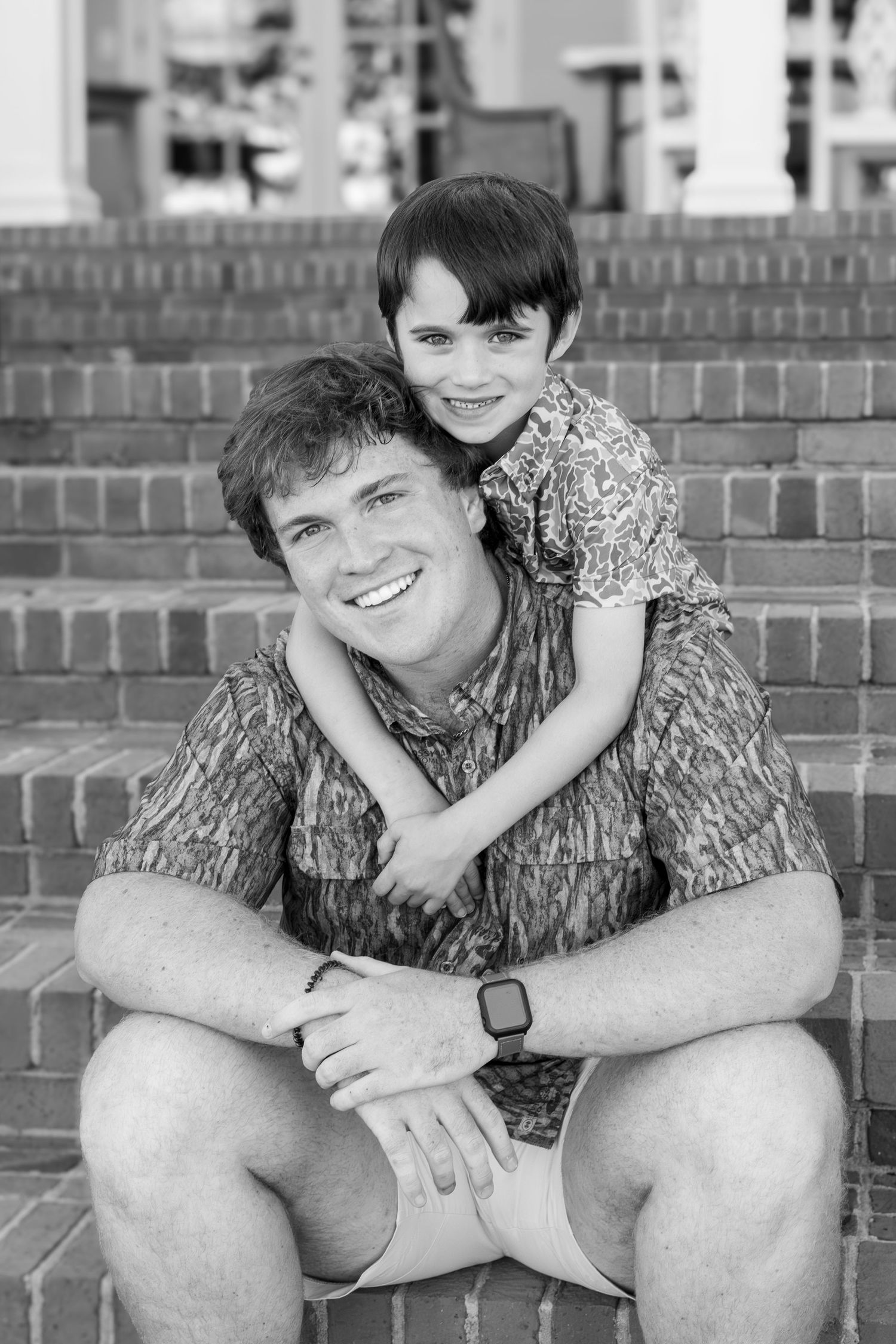 Black and white photo of a smiling man sitting on brick steps with a young boy hugging him from behind.