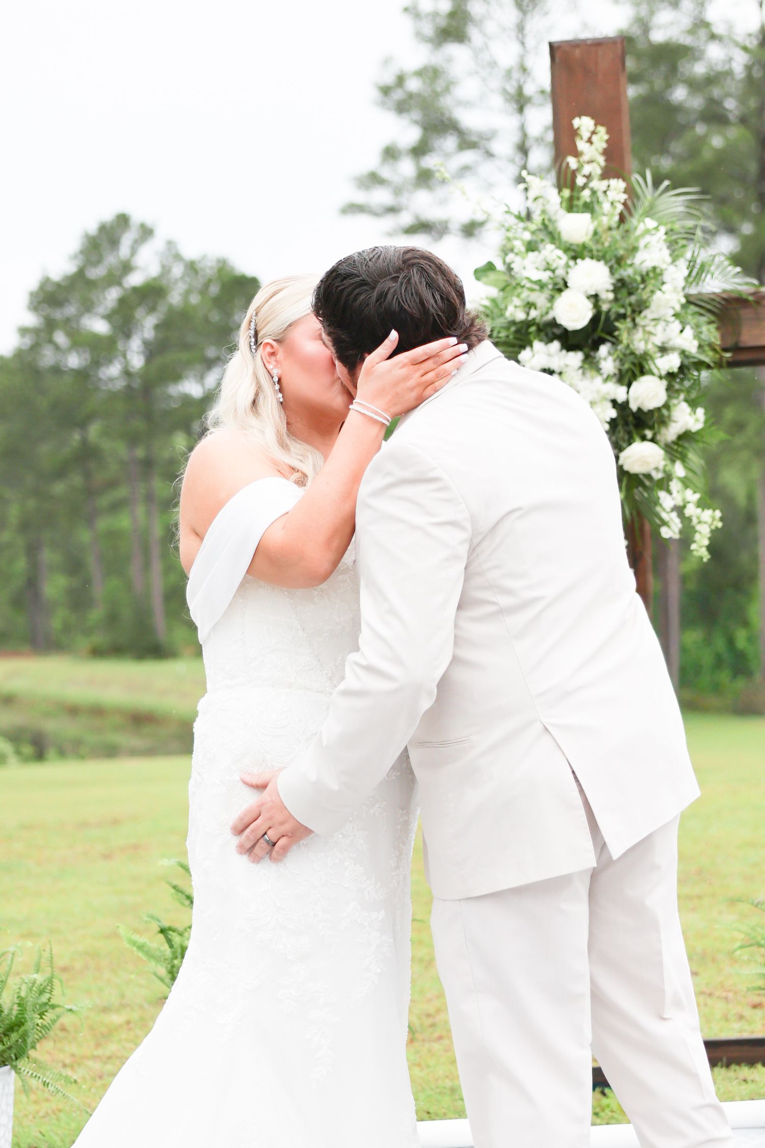 Bride and groom share first kiss at outdoor wedding altar adorned with white floral arrangements.