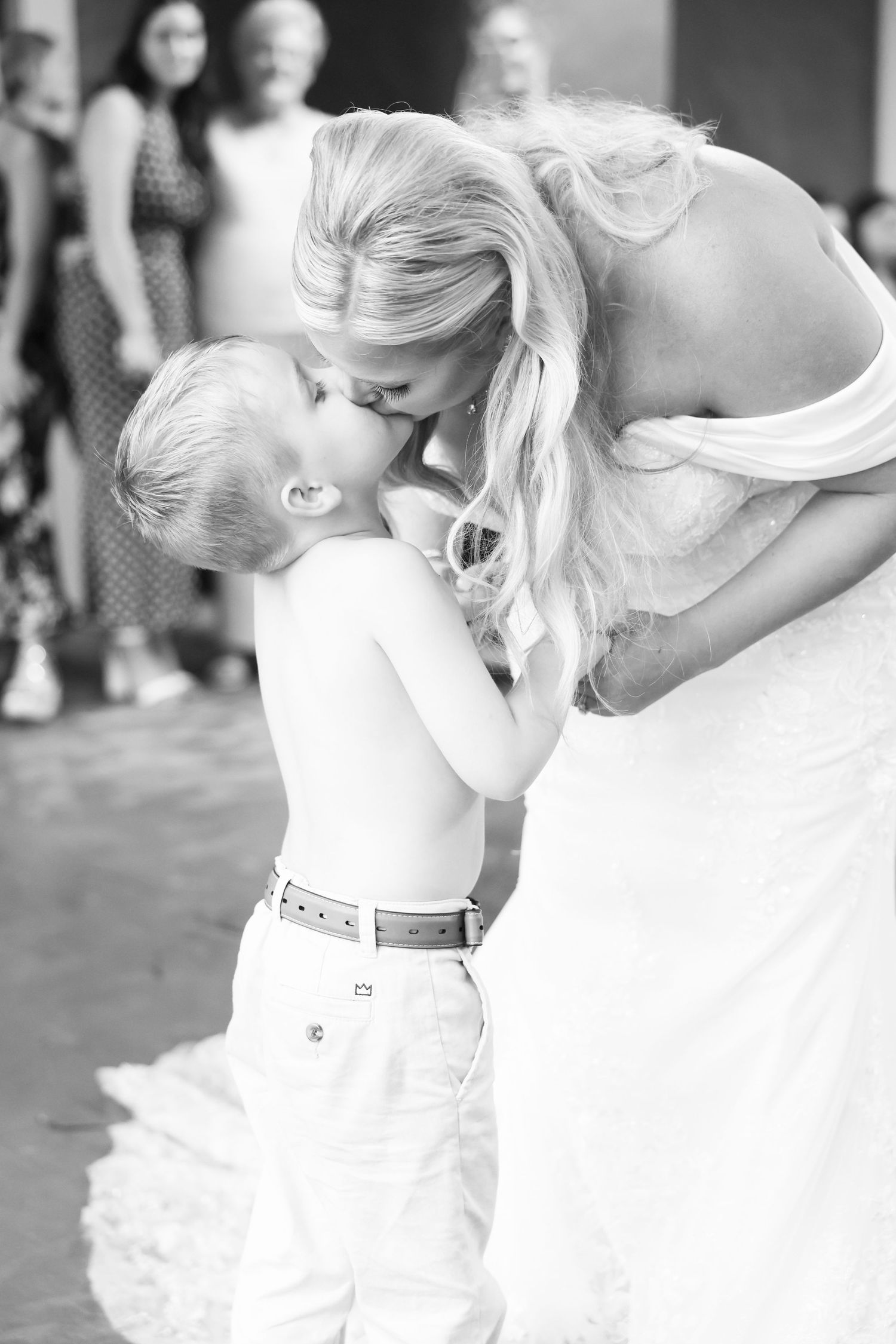 Black and white photo of a bride kissing a young boy on the forehead at a wedding reception.