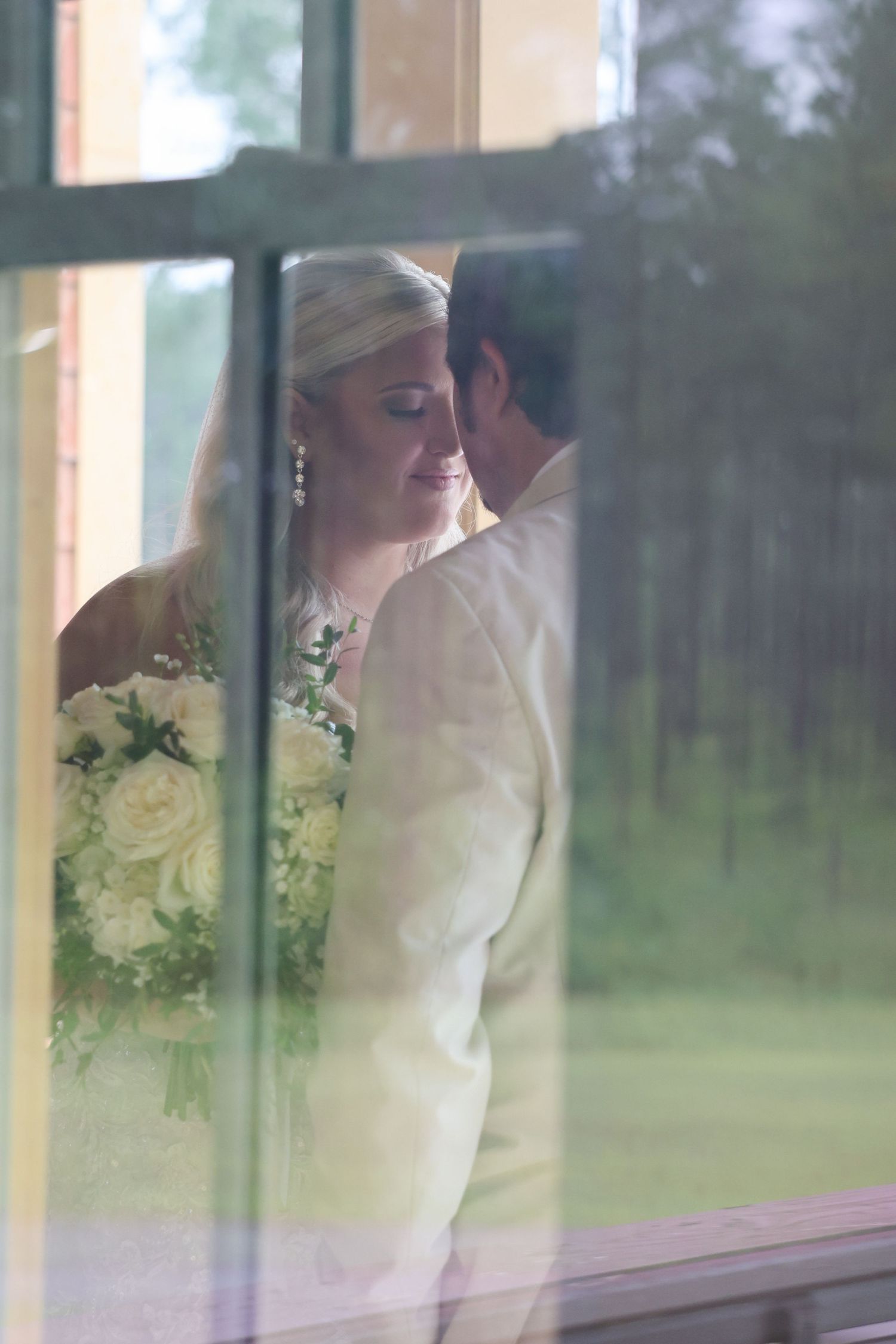 Bride and groom share a romantic moment by a window, bride holding white bouquet in elegant wedding attire.