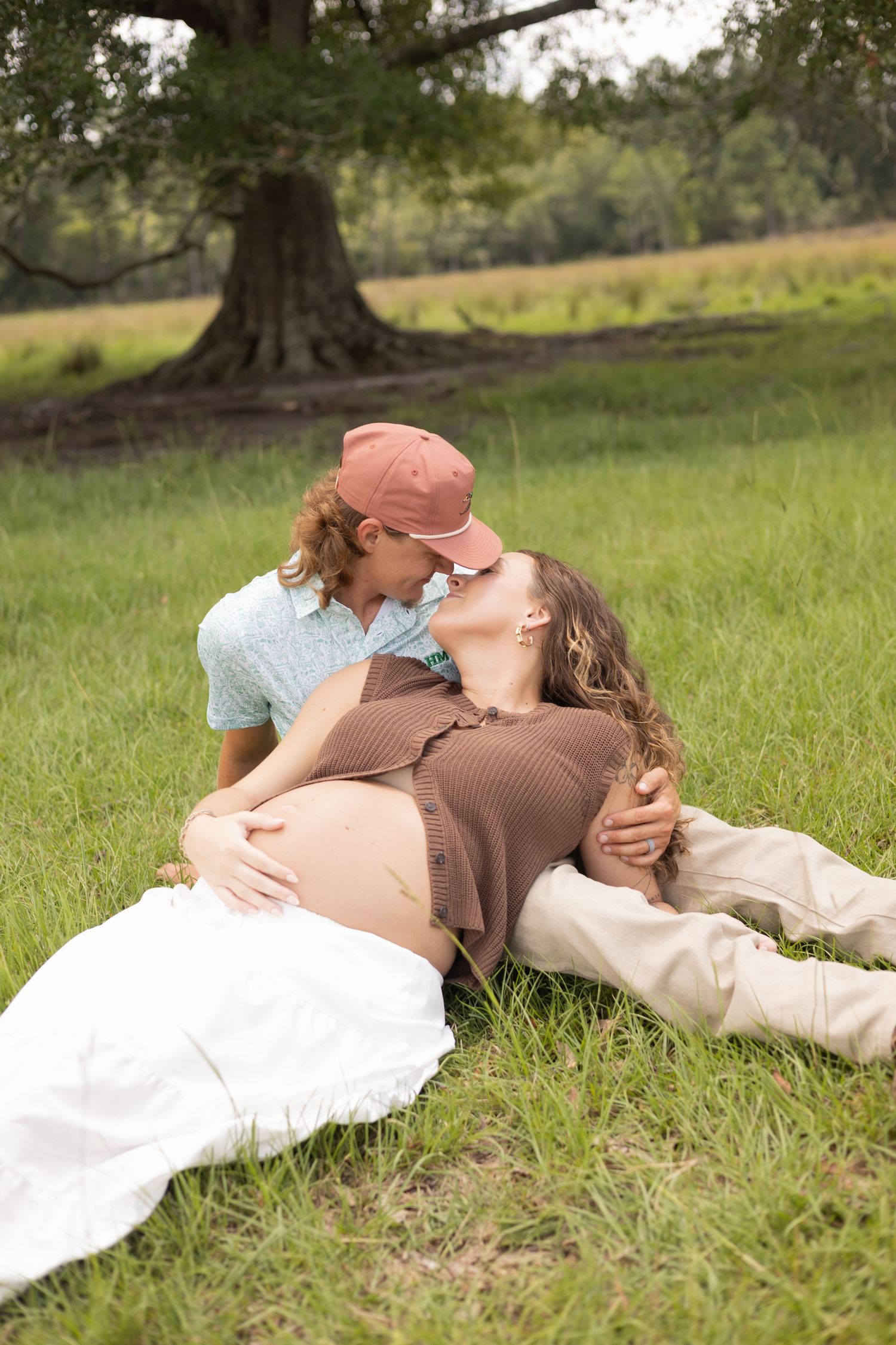 Pregnant couple sharing a kiss while relaxing on grass beneath a large tree in a lush green park.