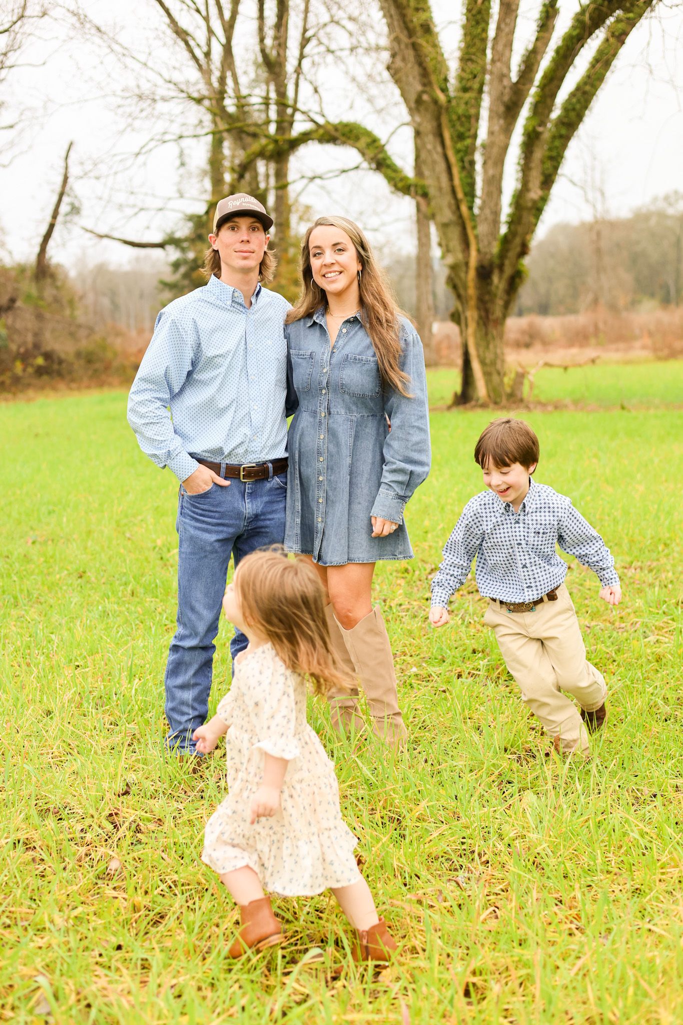 A family poses together while children run and play in a green field with tall trees.