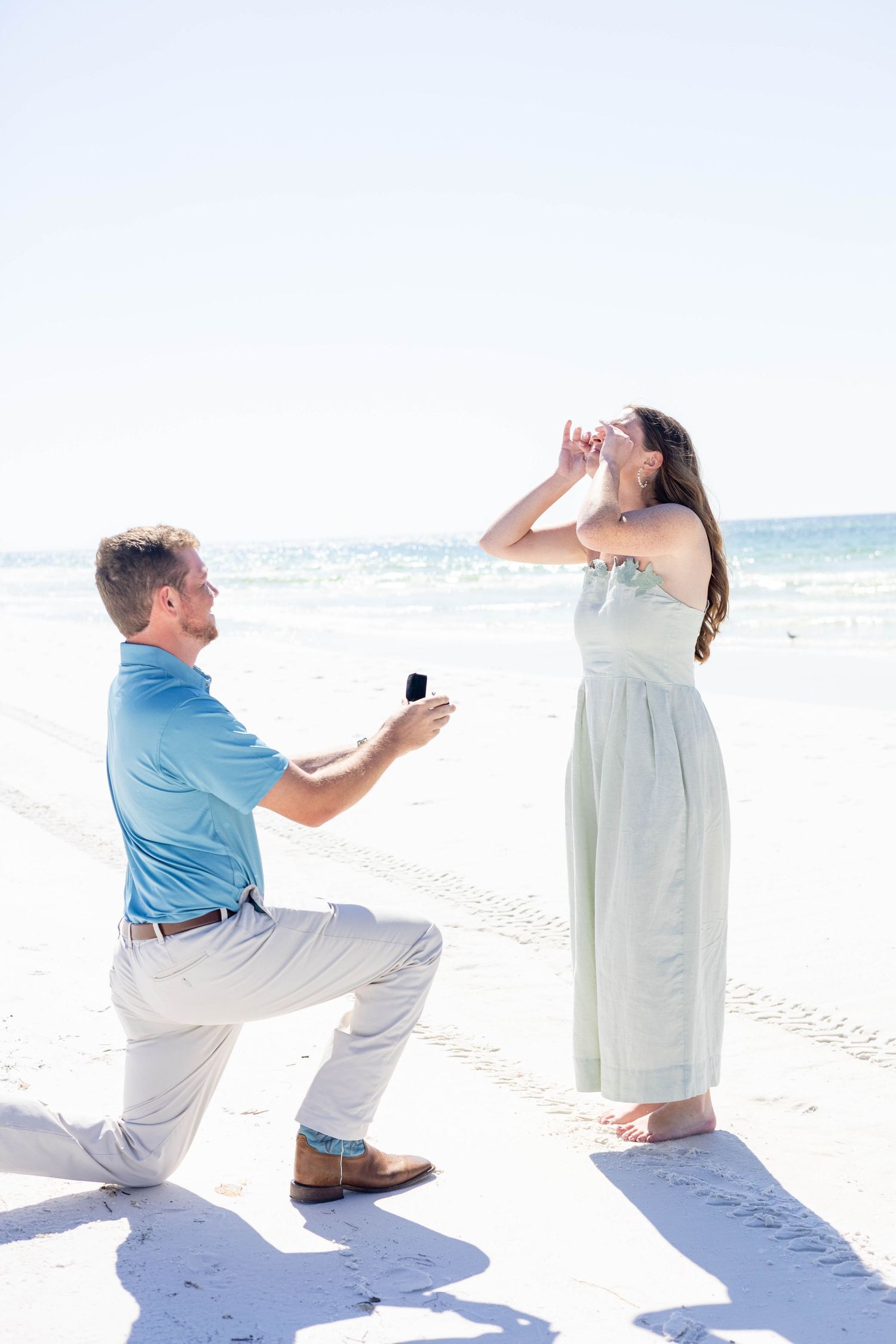 A romantic beach proposal scene with someone kneeling in the sand presenting a ring.