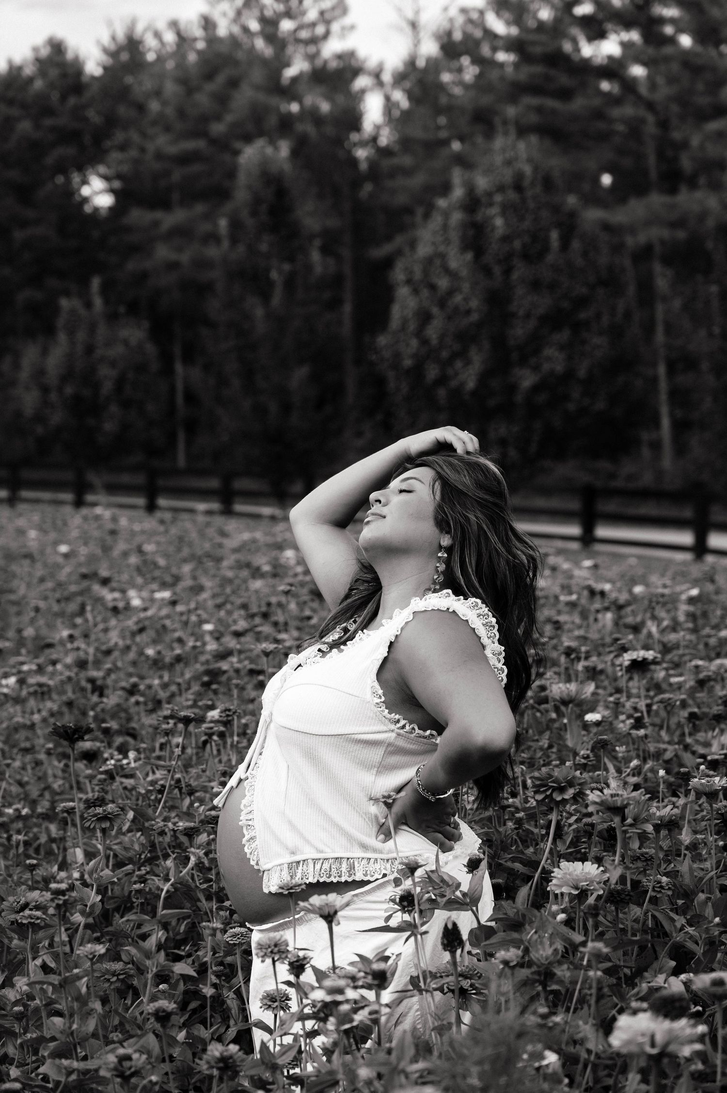 Black and white photo of a pregnant woman in a white dress posing gracefully in a flower field with trees in background.