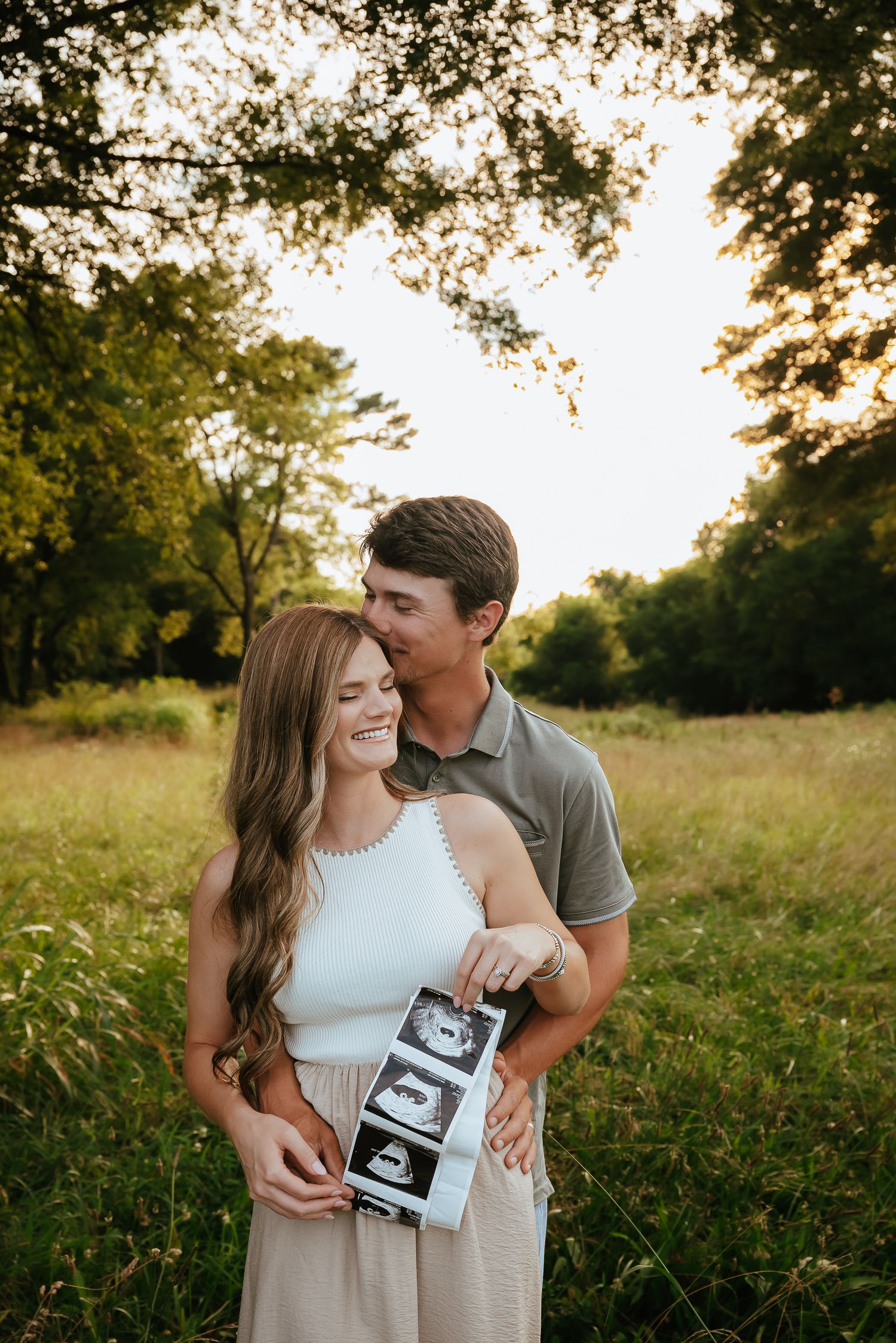 Outdoor pregnancy announcement photoshoot in a sunny field with ultrasound photos.
