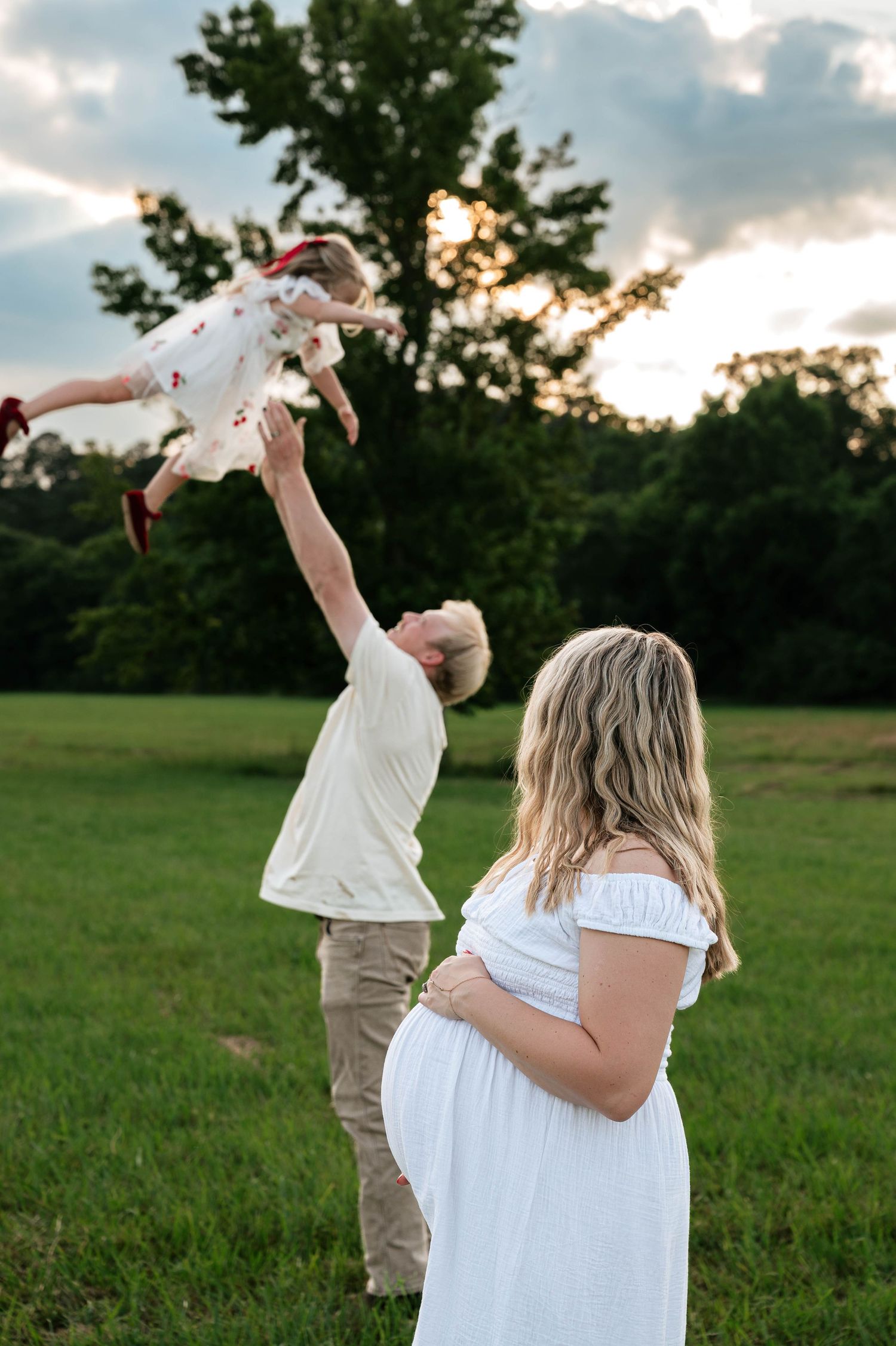 Pregnant woman in white dress watches partner toss young child in air during outdoor maternity photo session at sunset.