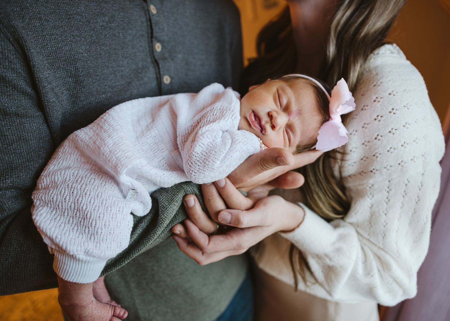 Newborn baby in pink bow headband sleeping peacefully while being held by parents in a tender moment.
